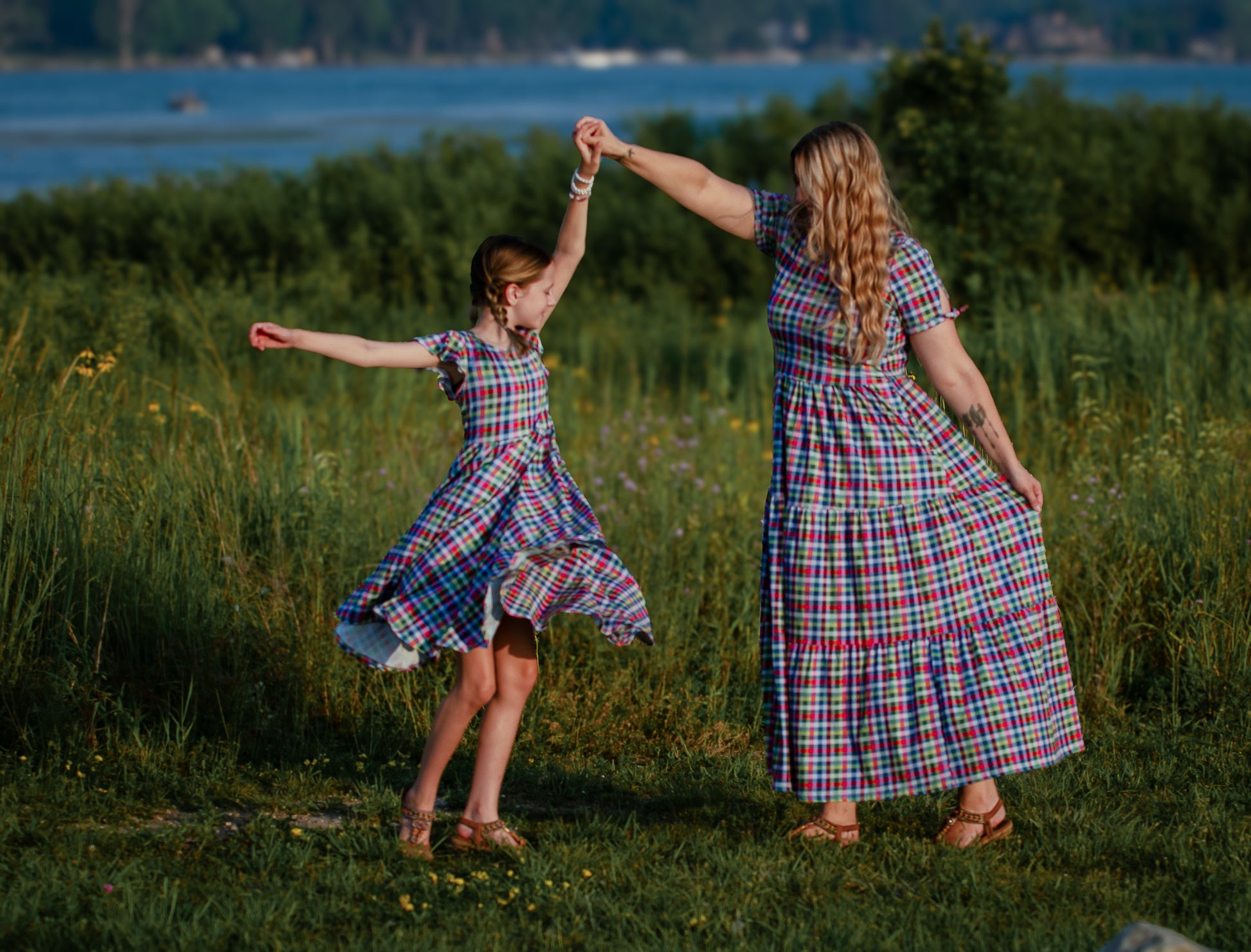 Mother and daughter in plaid modest dresses dancing in a field with a lake in the background