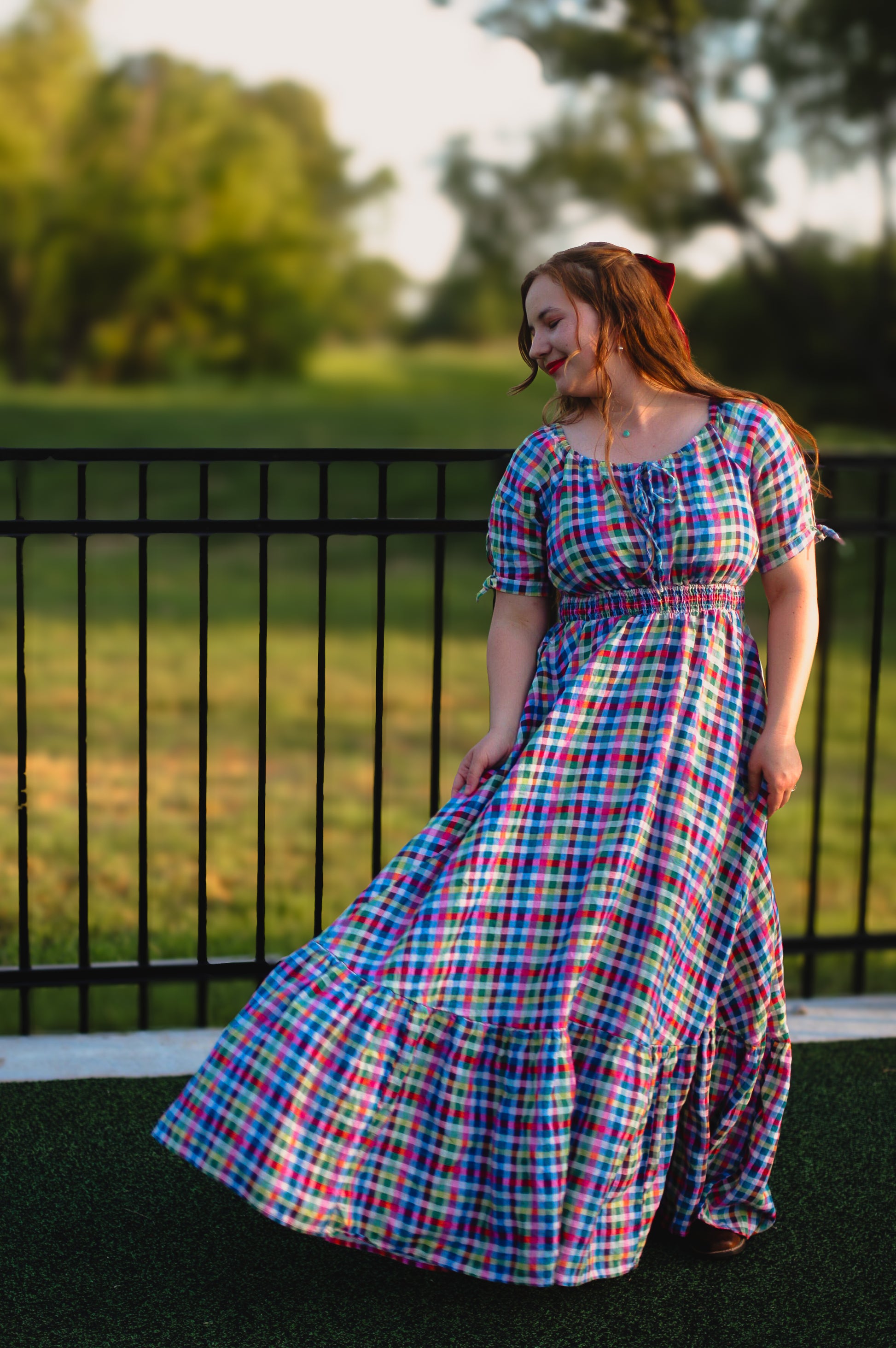 Woman wearing a plaid modest nursing dress standing outdoors with trees and a fence in the background