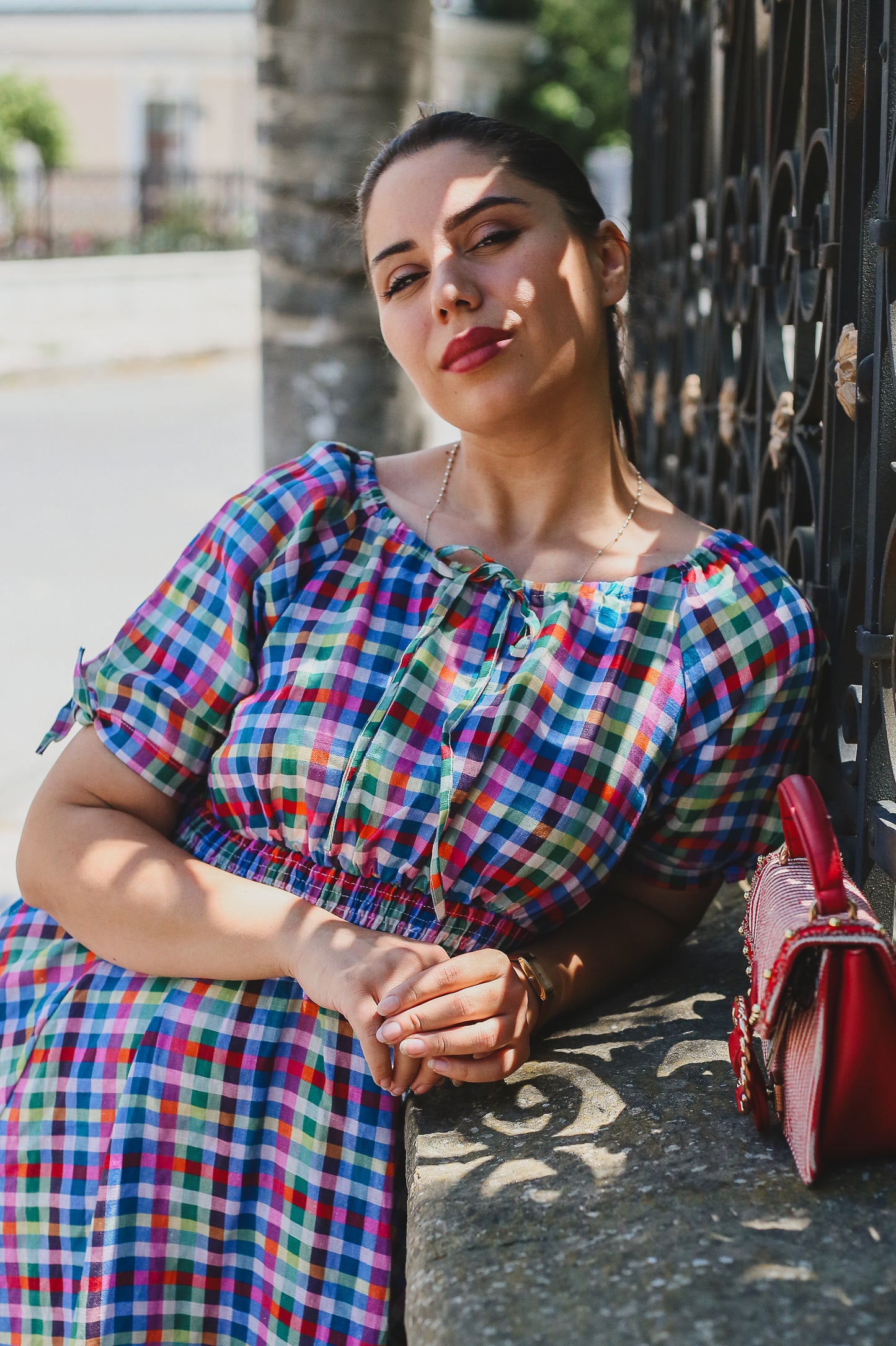 Woman in a colorful checkered modest nursing dress sitting on a stone wall with a red handbag beside her.