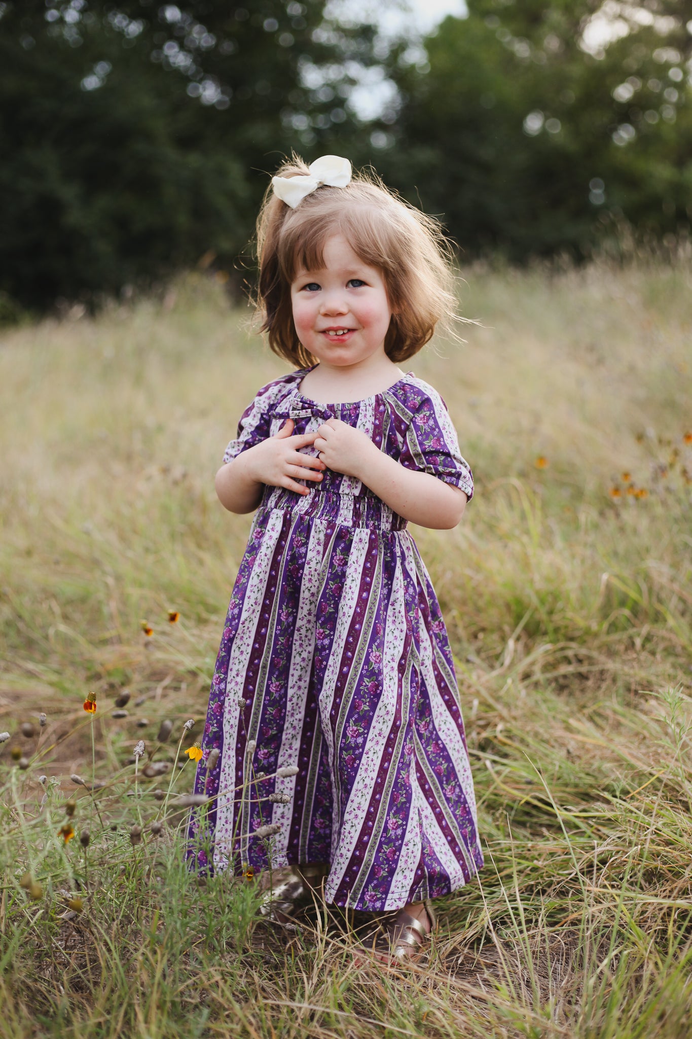 Young girl in a purple dress standing in a grassy field with trees in the background