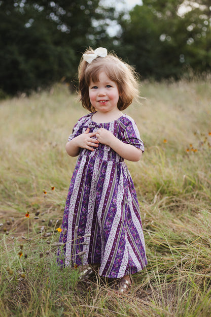 Young girl in a purple dress standing in a grassy field with trees in the background