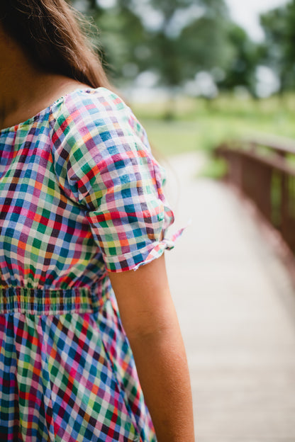 Person wearing a colorful checkered modest nursing dress standing on a wooden bridge with a blurred natural background