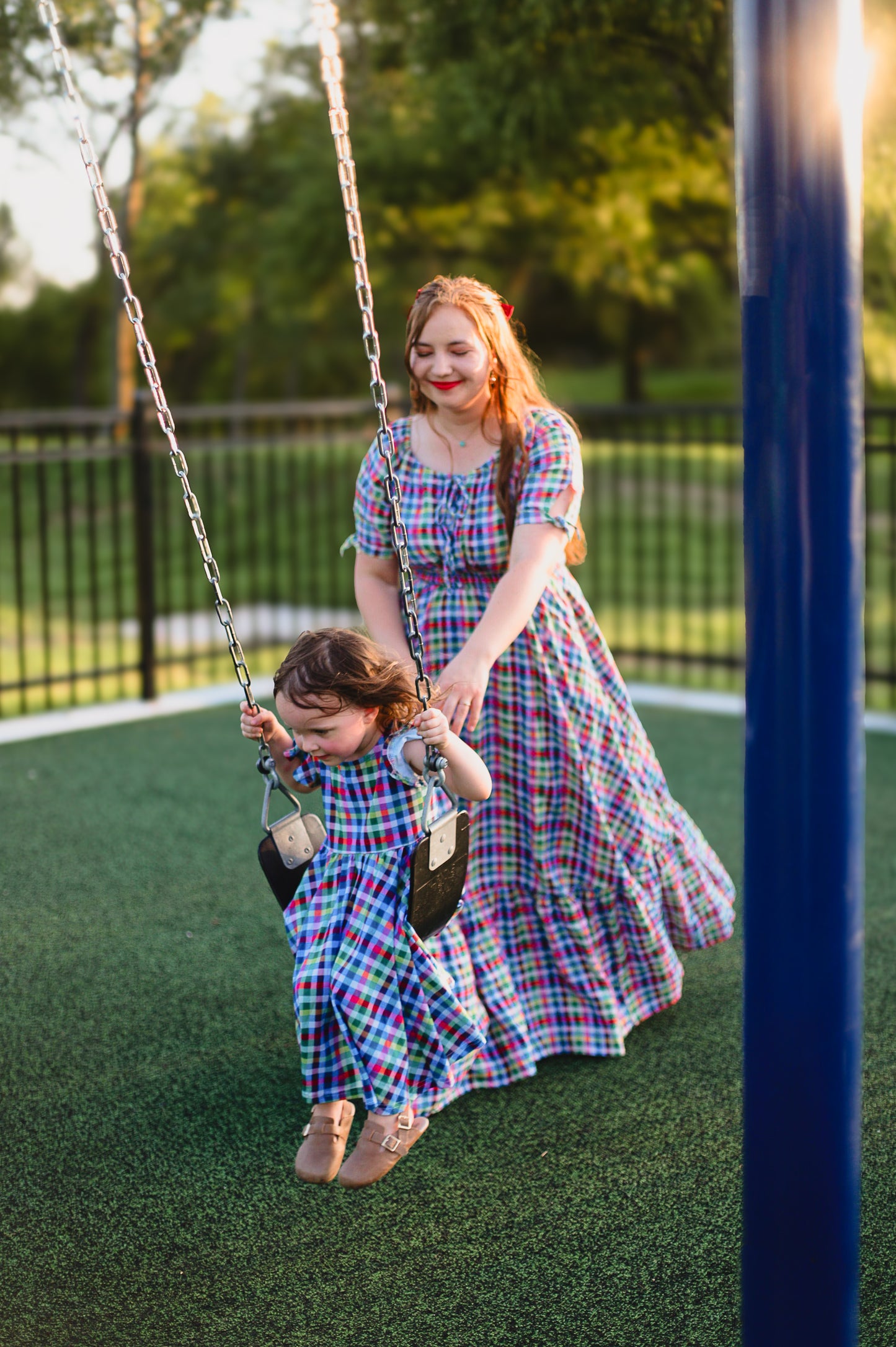 Mother and daughter in plaid modest dresses swinging a child on a playground swing set.