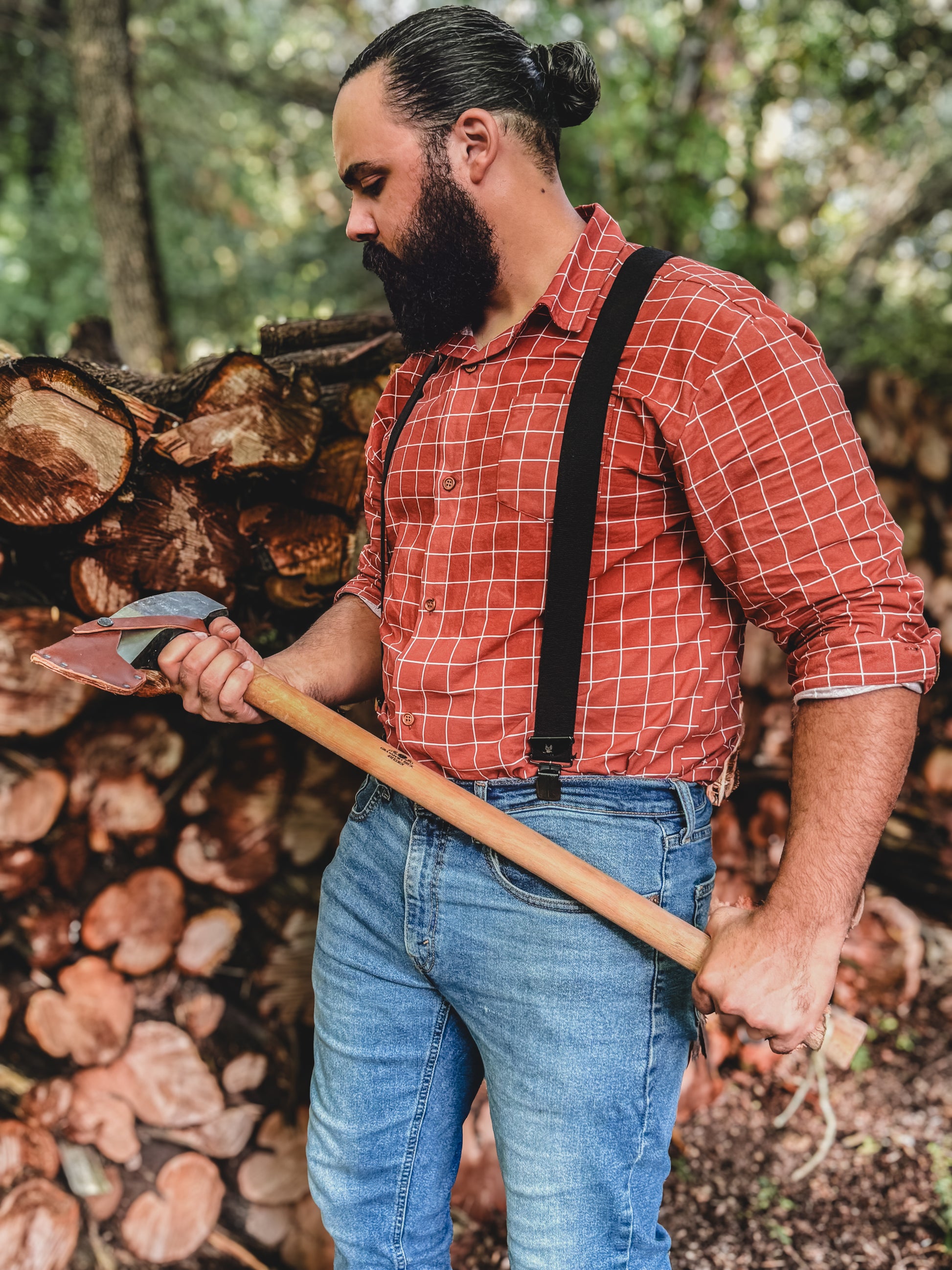Man in a red plaid shirt and suspenders holding an axe in front of stacked logs.
