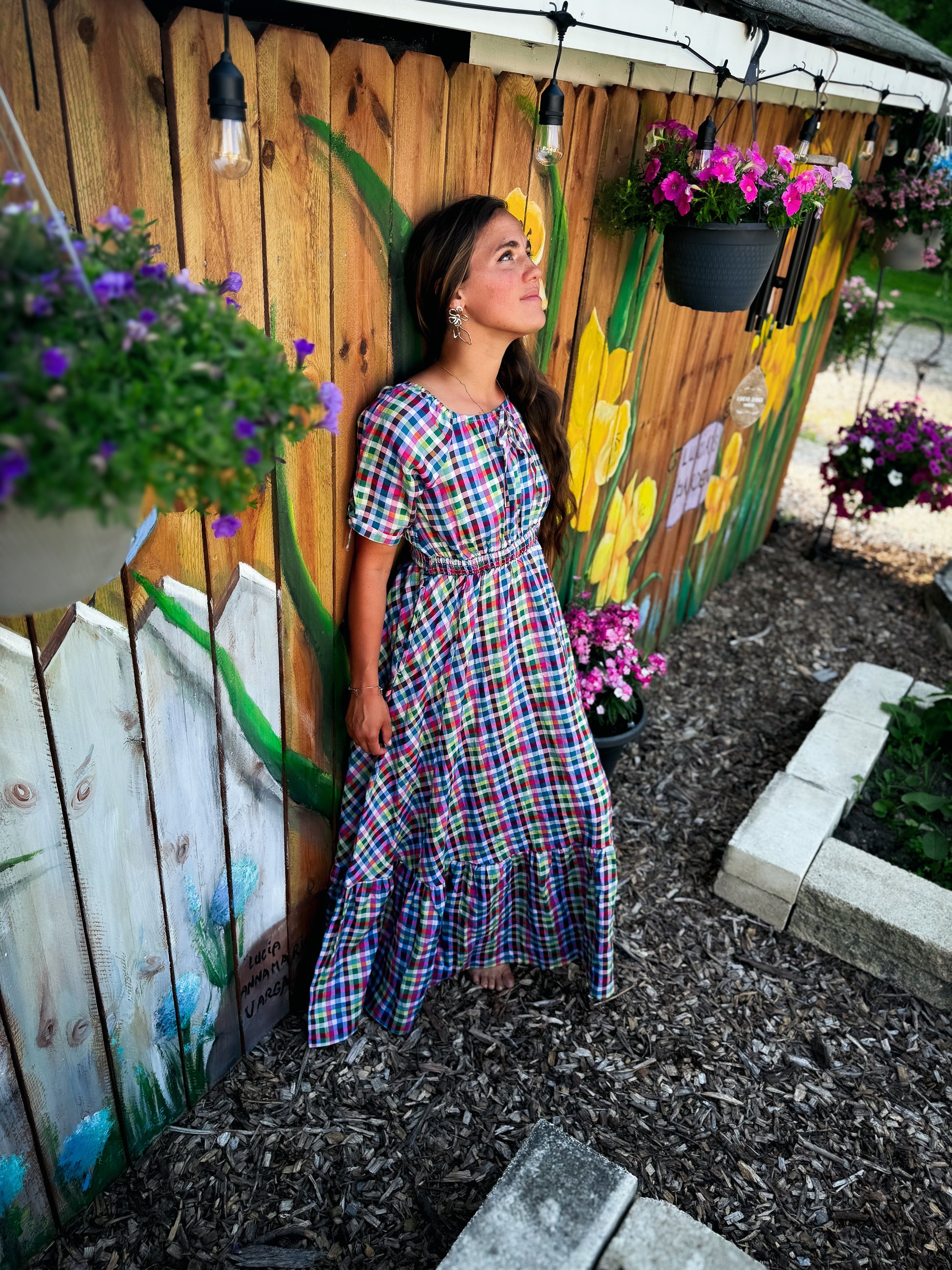 Woman in a plaid modest nursing dress standing in a garden with flowers and a wooden fence.
