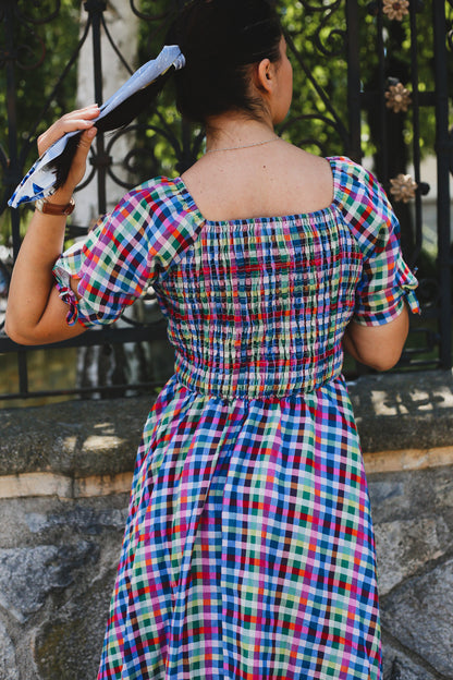 Woman wearing a colorful checkered modest nursing dress standing against a stone wall with a metal gate.