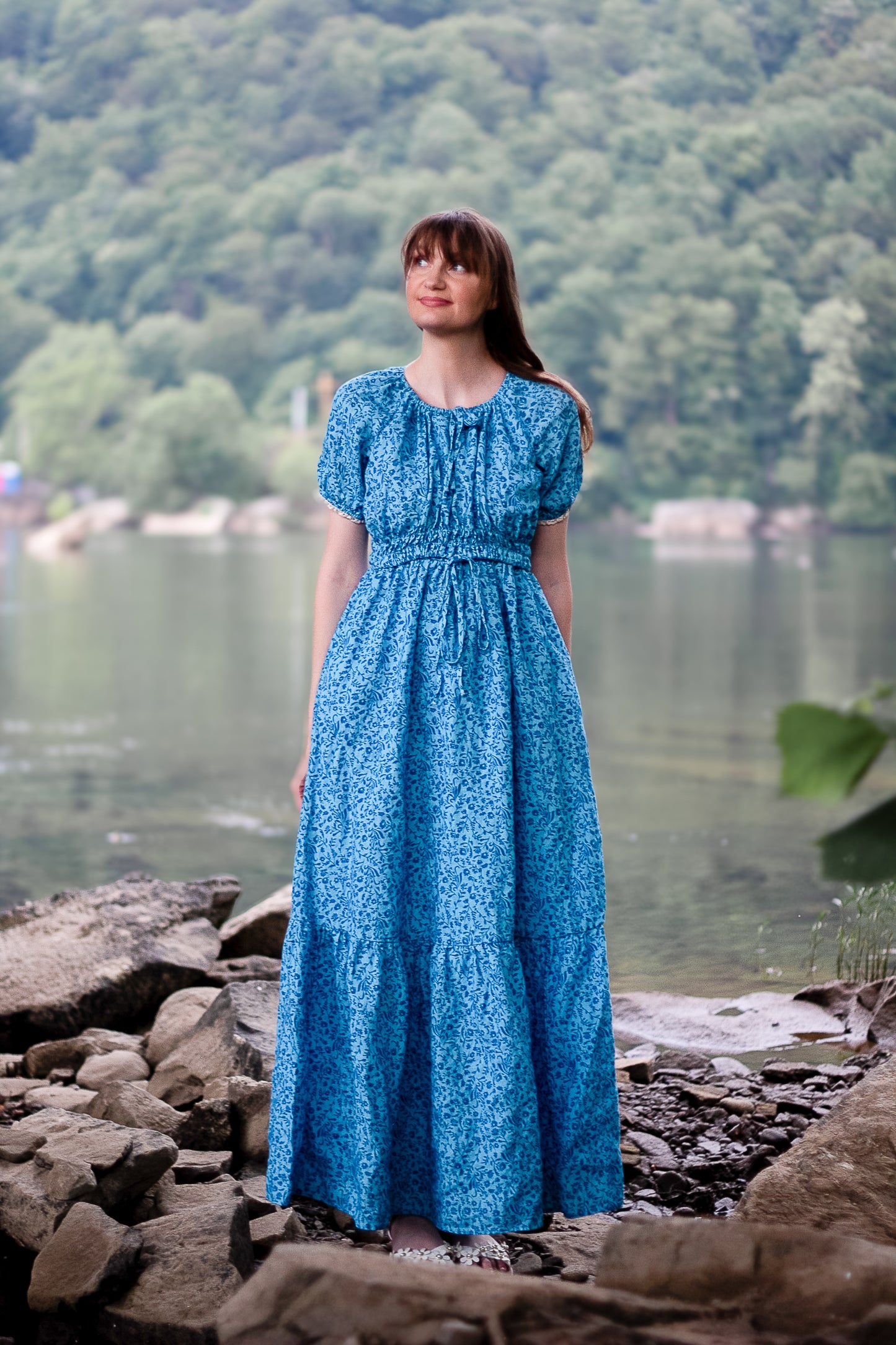 Woman in a blue modest nursing dress standing by a lake with mountains in the background