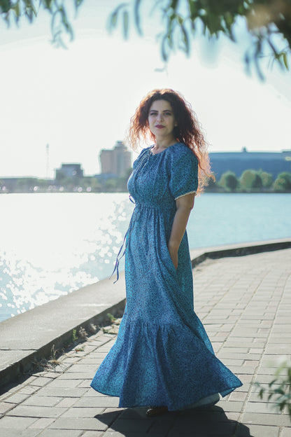 Woman in a blue modest nursing dress standing by a waterfront with buildings in the background