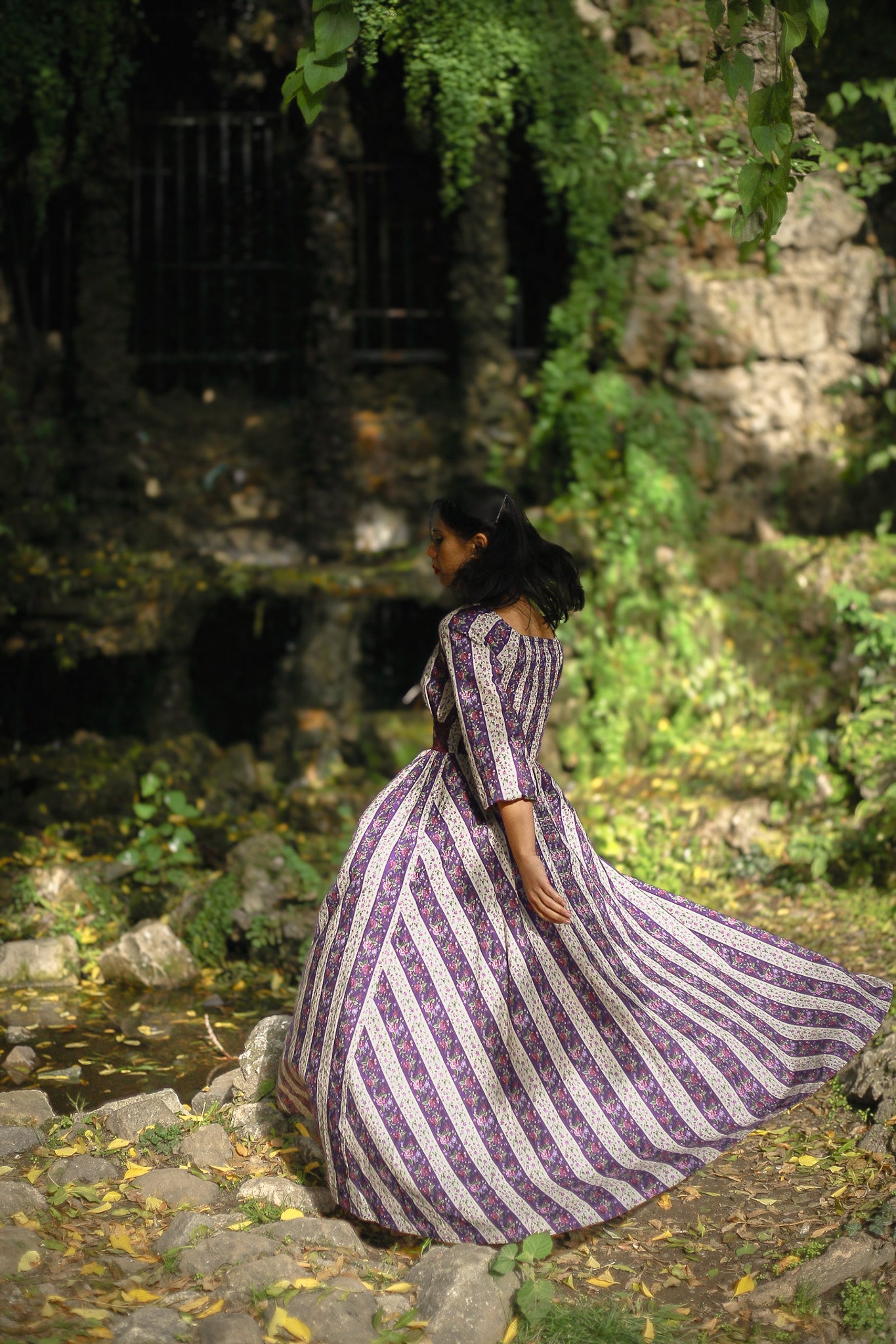 Woman in a modest nursing purple and white striped dress standing in a natural setting with greenery and rocks.
