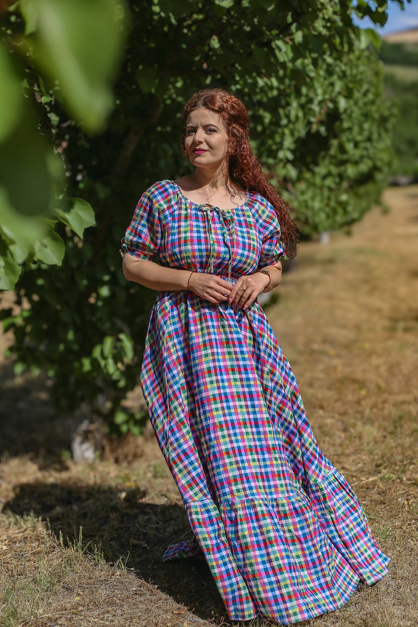 Woman in a colorful checkered modest nursing dress standing in a field with trees in the background