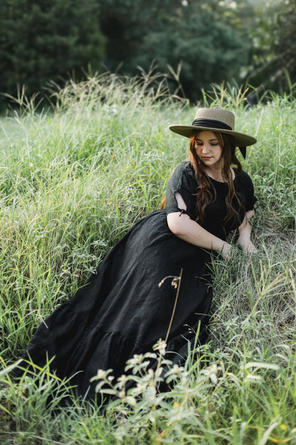 Woman in a black modest nursing dress and hat sitting in a grassy field