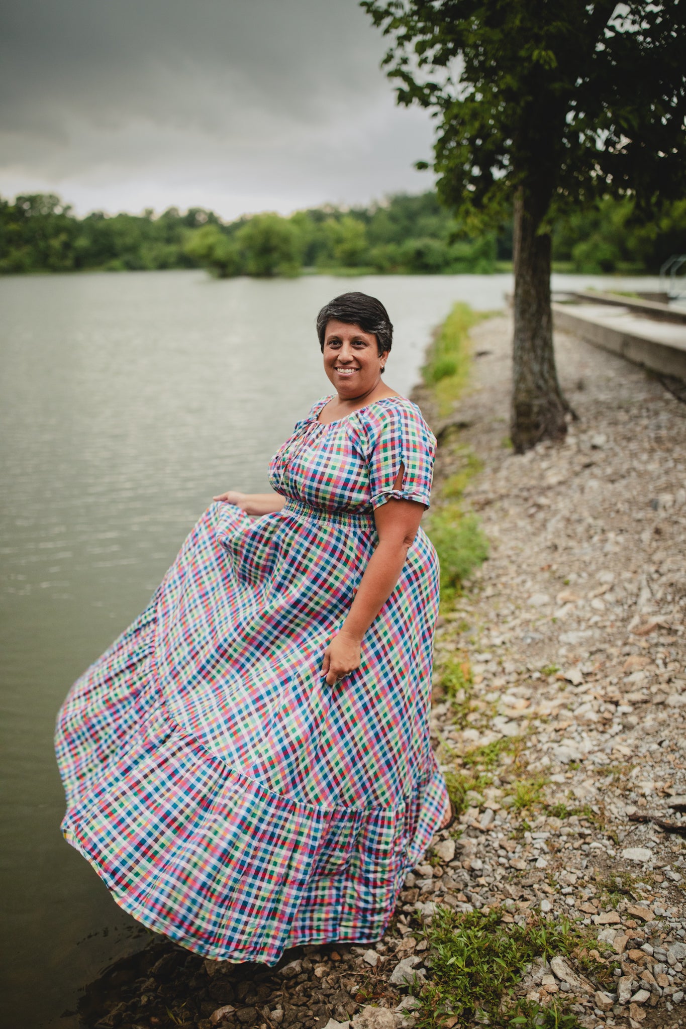 Woman in a plaid modest nursing dress standing by a lake with trees in the background