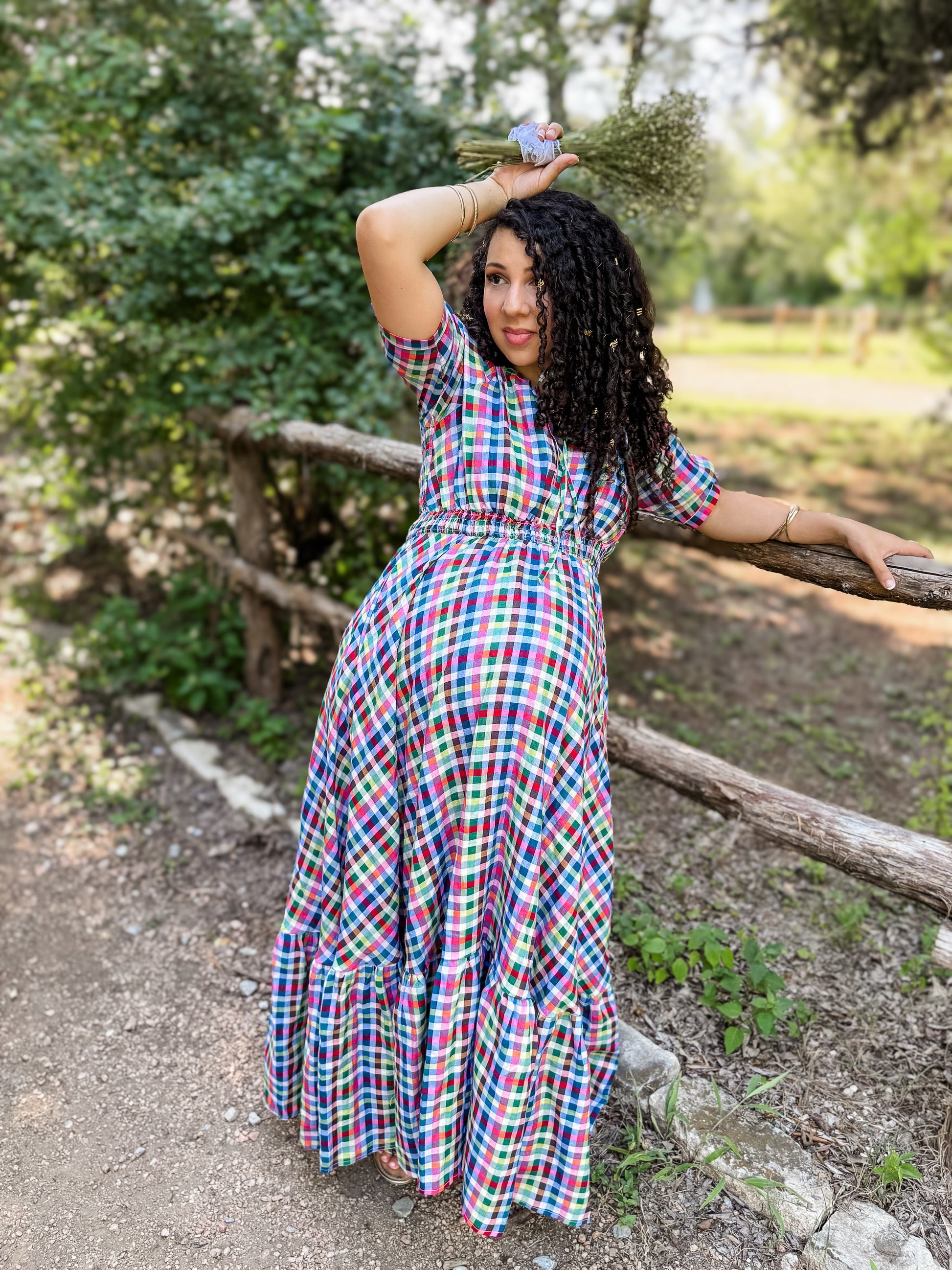Woman in a colorful plaid modest nursing dress standing outdoors near a wooden fence with greenery in the background.