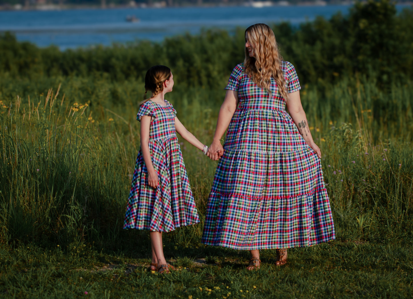 Mother and daughter in matching plaid modest dresses standing in a grassy field by a body of water.
