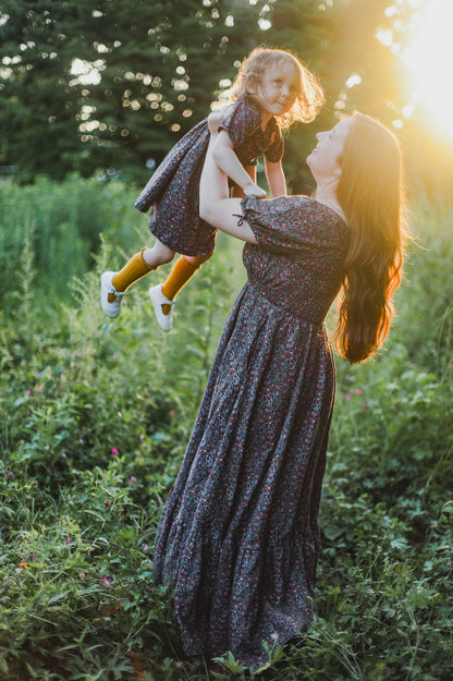 Woman holding a child in a field with sunlight filtering through wearing a modest nursing dress