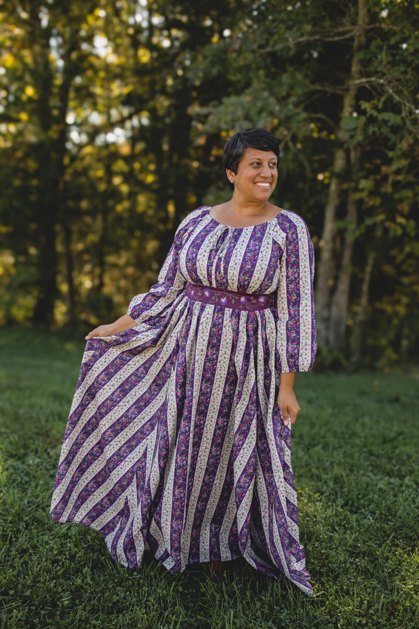 Woman wearing a modest nursing purple and white striped dress standing in a grassy area with trees.