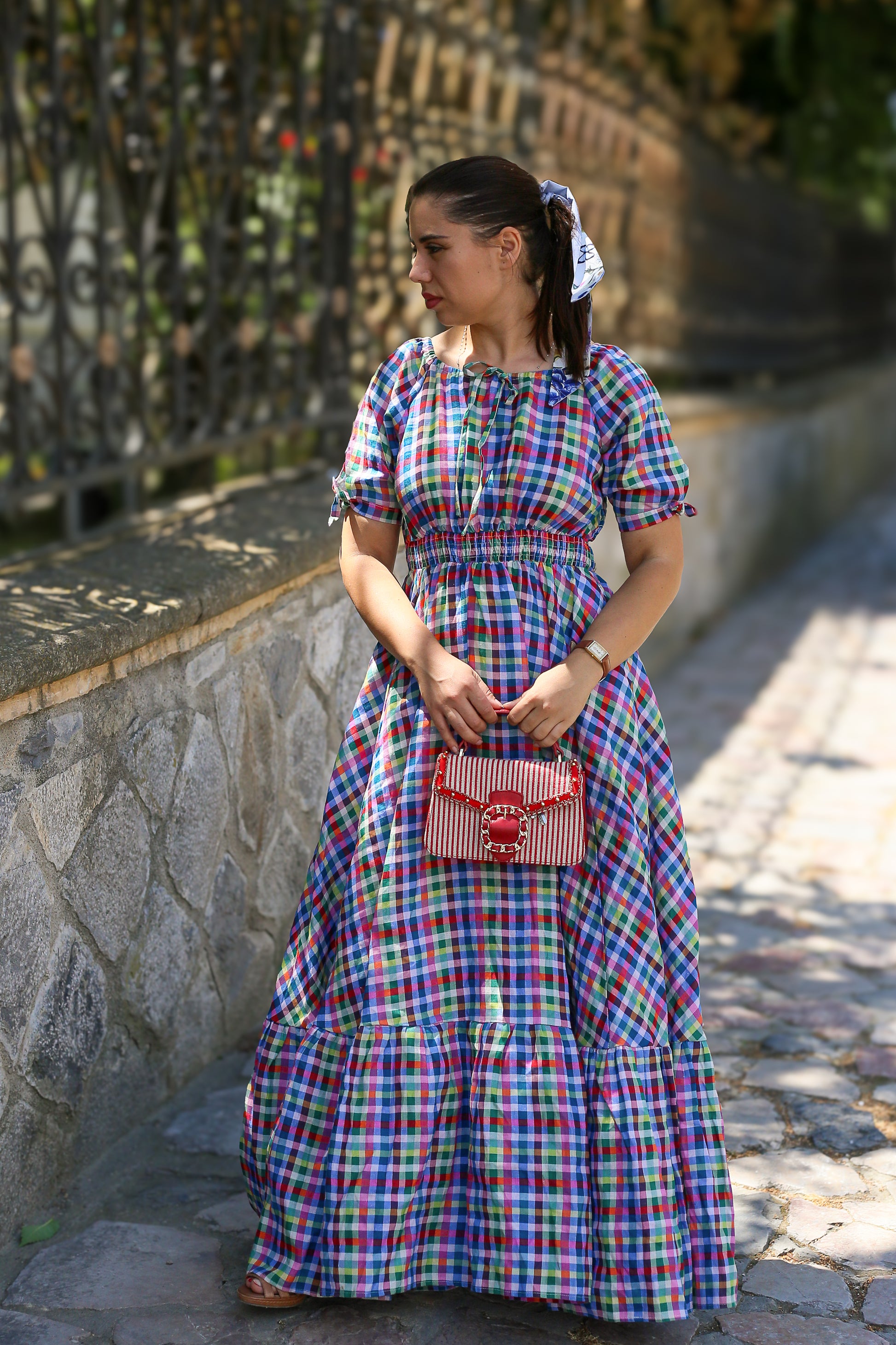 Woman in a colorful plaid modest nursing dress holding a red handbag on a stone path.
