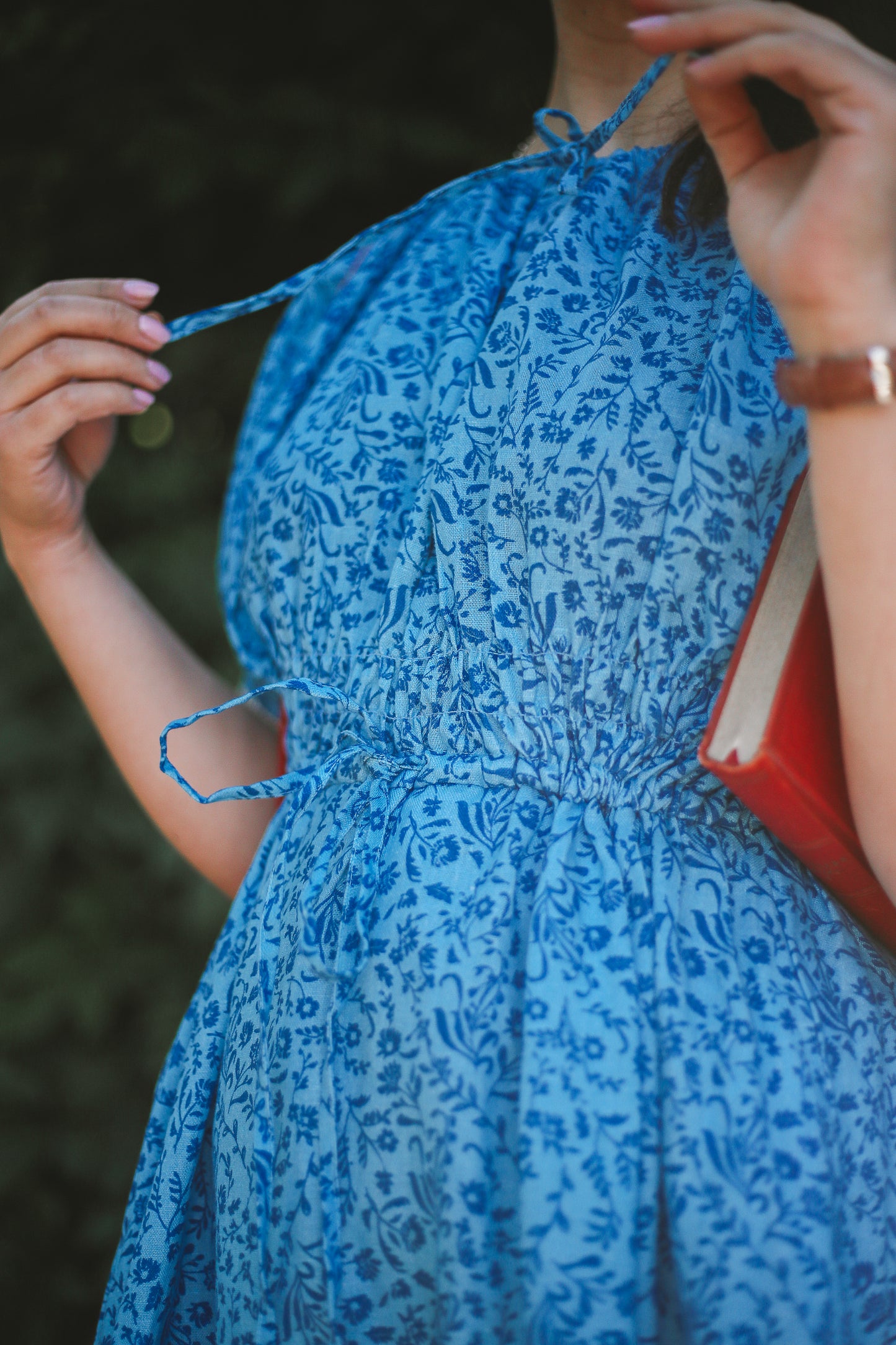 Woman wearing a blue floral modest nursing dress holding a red book against a blurred natural background