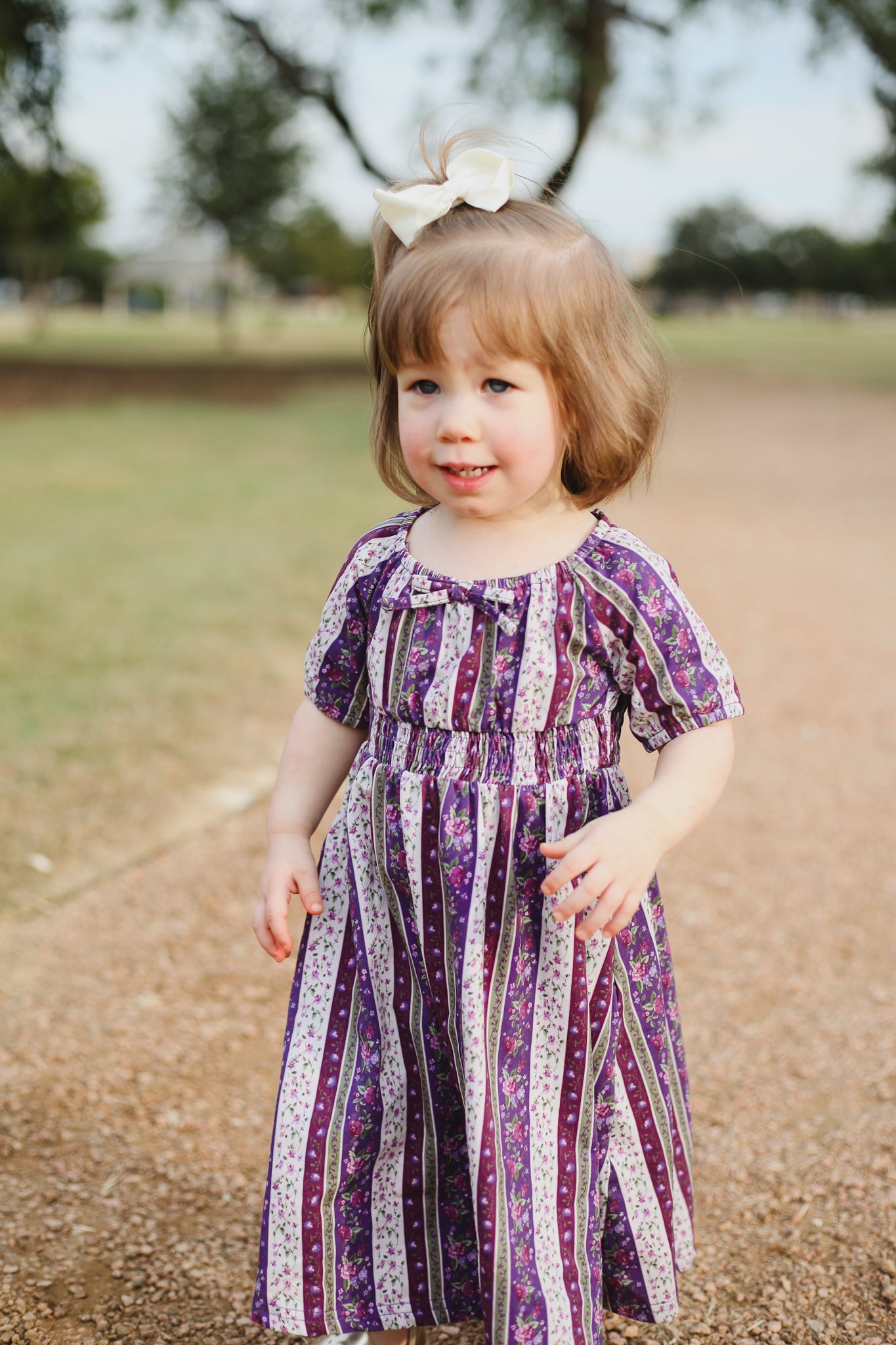 Young girl in a purple patterned dress standing on a dirt path with trees in the background