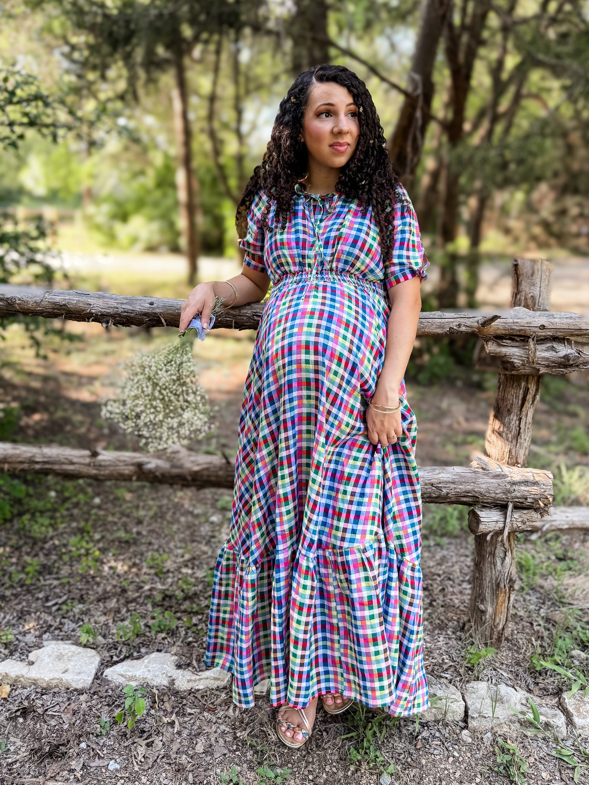 Woman in a colorful modest nursing dress standing outdoors by a wooden fence with trees in the background