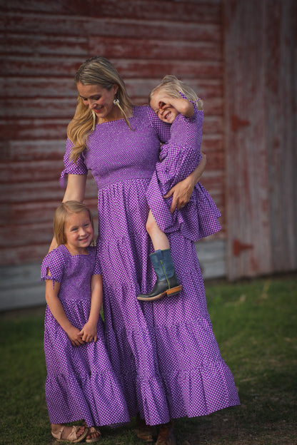A woman wearing a purple, gingham, five-tiered maxi modest nursing dress with her two daughters in matching dresses