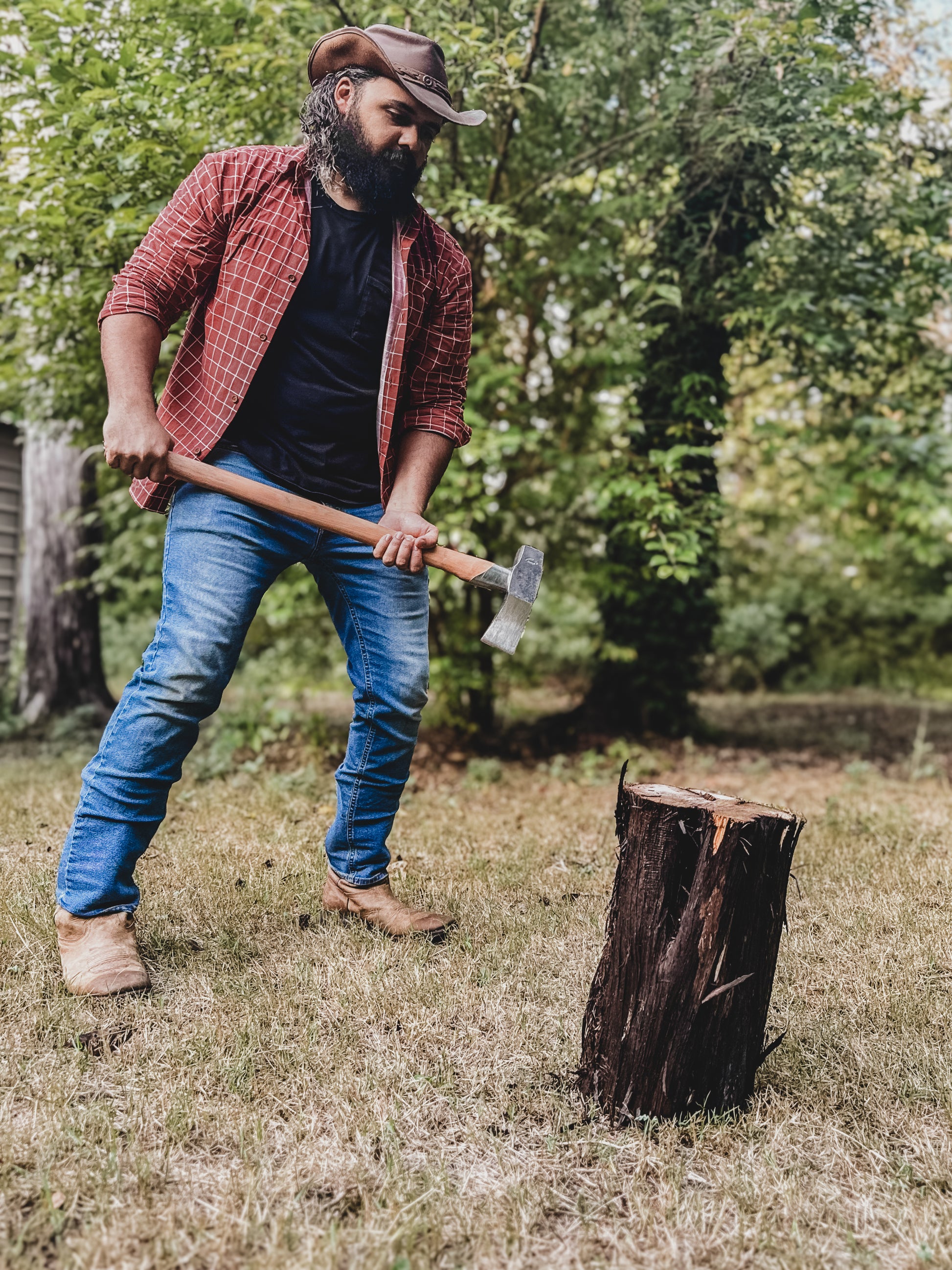 Man in a plaid shirt and jeans holding an axe next to a wooden stump in a forest setting