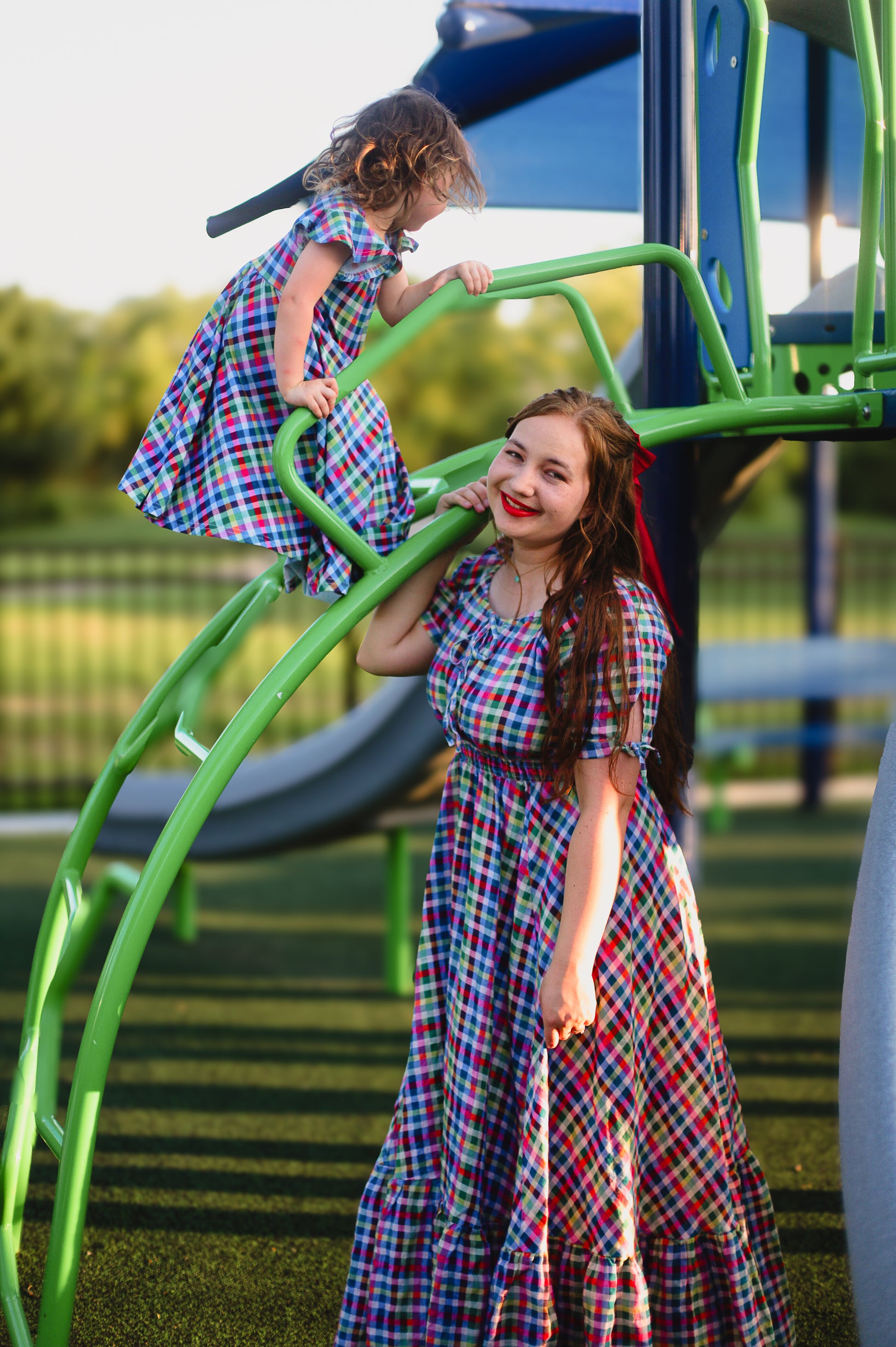 Mother and daughter in plaid modest dresses on a playground with green equipment.