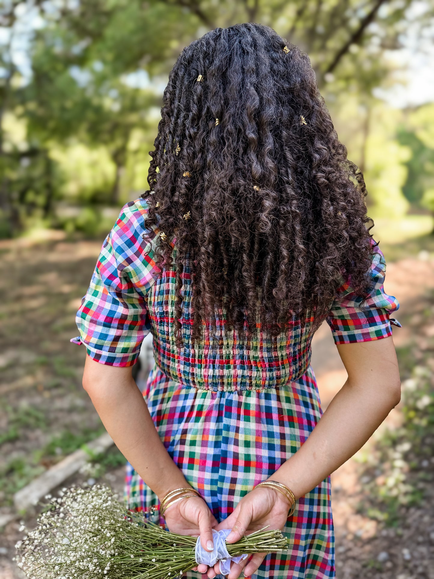 Person with long, braided hair wearing a colorful modest nursing dress holding flowers outdoors.