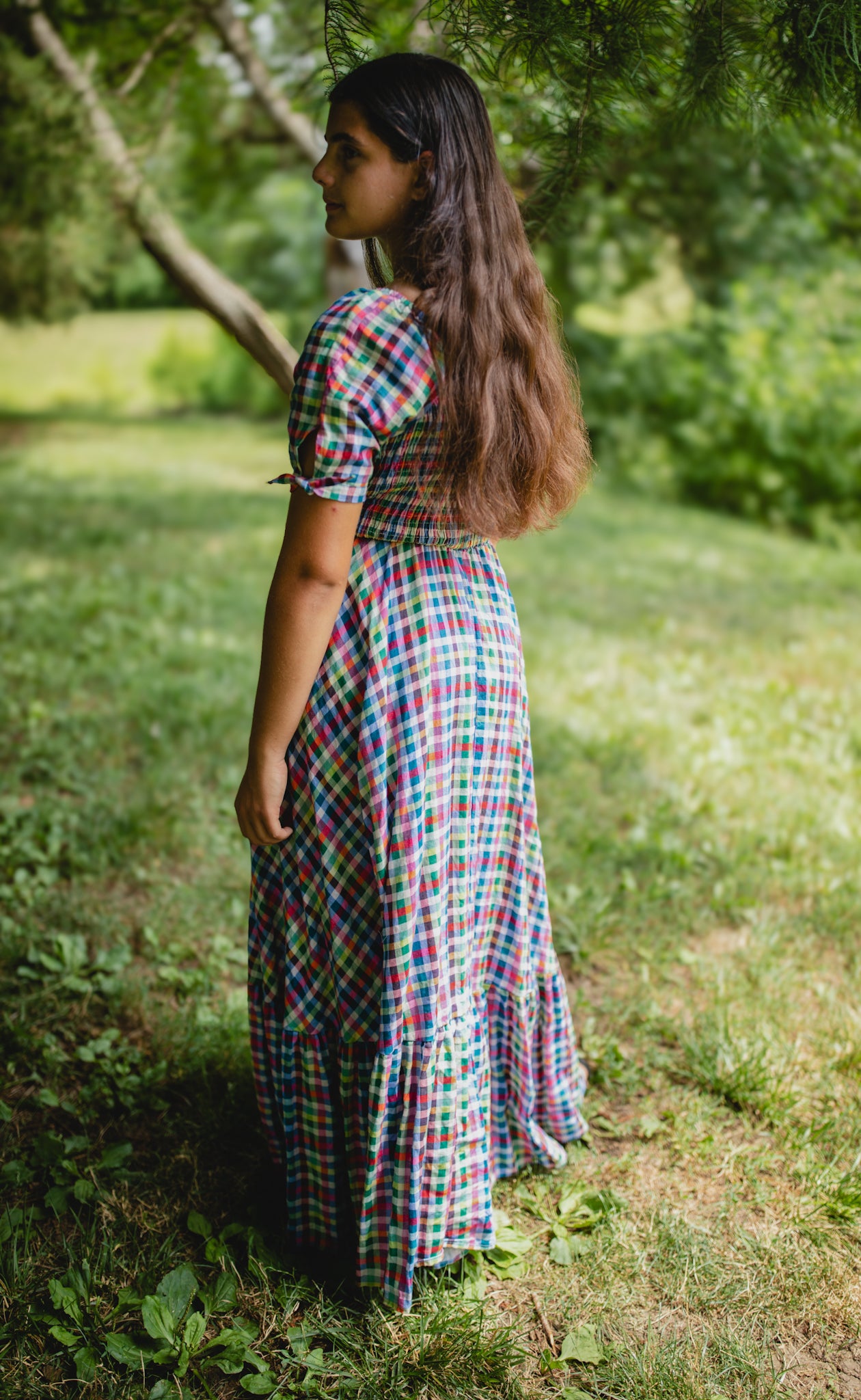 Woman in a plaid modest nursing dress standing in a grassy area with trees in the background