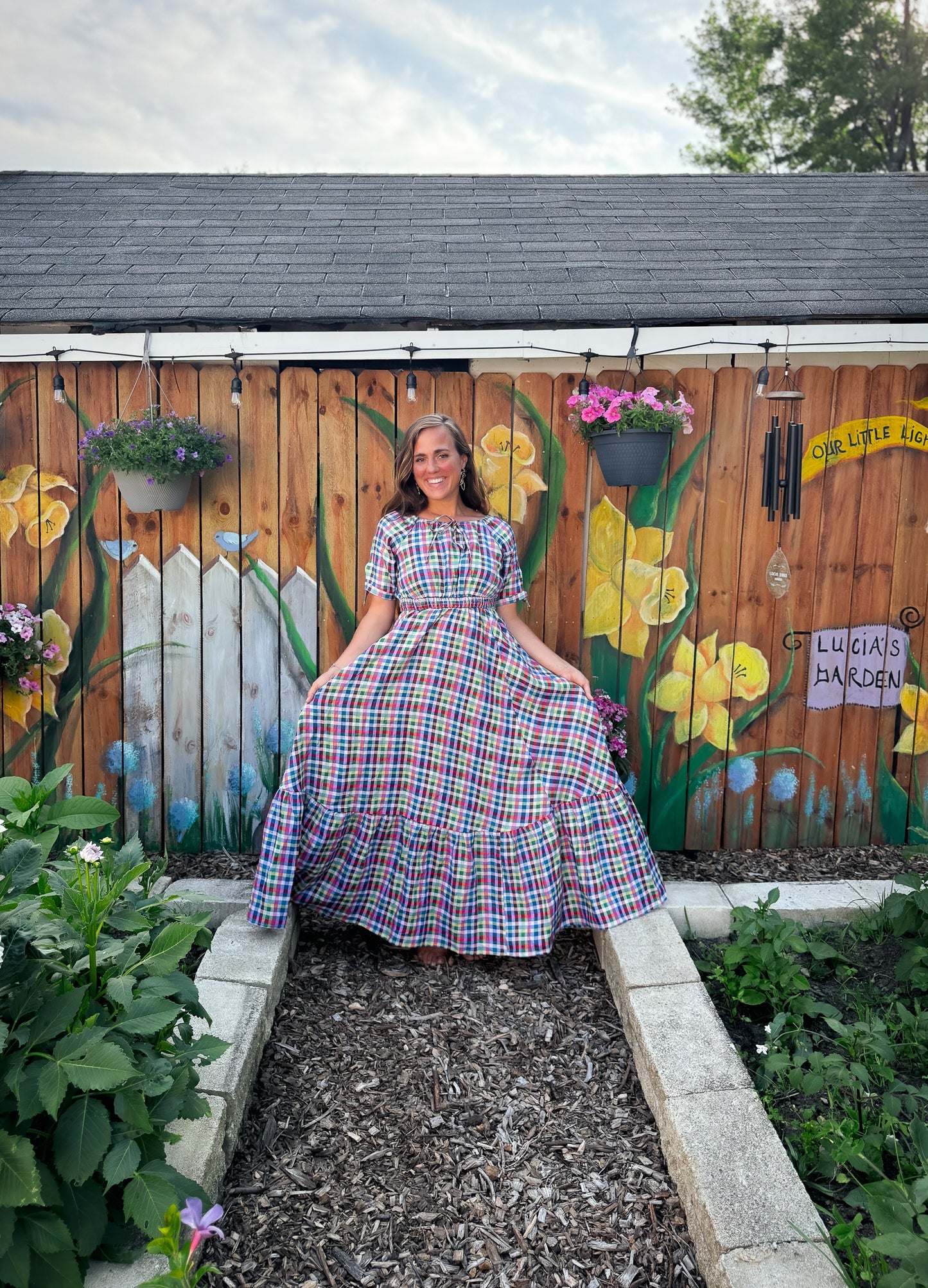 Woman in a plaid modest nursing dress standing in a garden with a wooden fence featuring floral artwork.