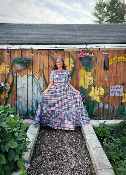 Woman in a plaid modest nursing dress standing in a garden with a wooden fence featuring floral artwork.