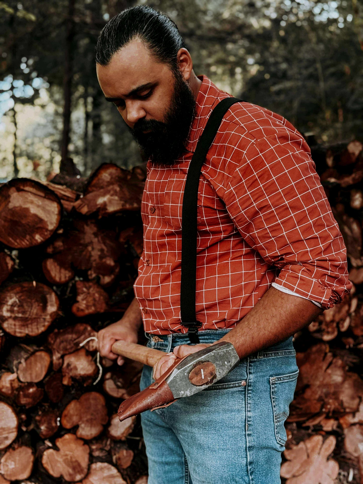 Man with a beard wearing a red plaid shirt and suspenders, holding a tool in front of stacked logs.