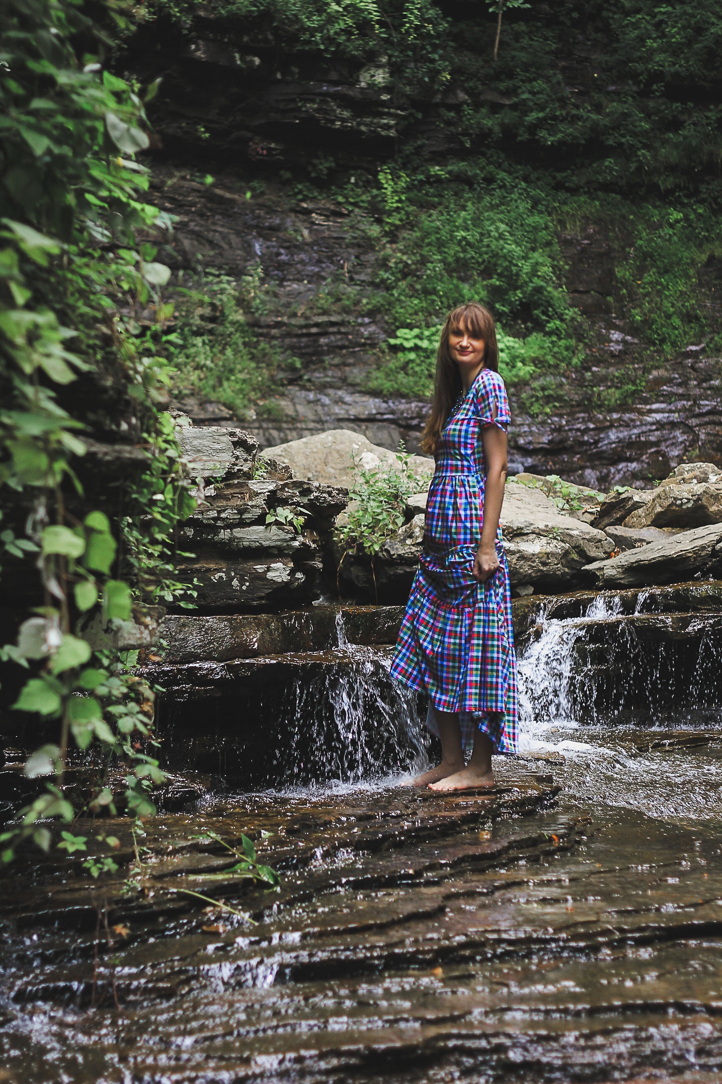 Woman in a plaid modest nursing dress standing by a small waterfall in a forest.
