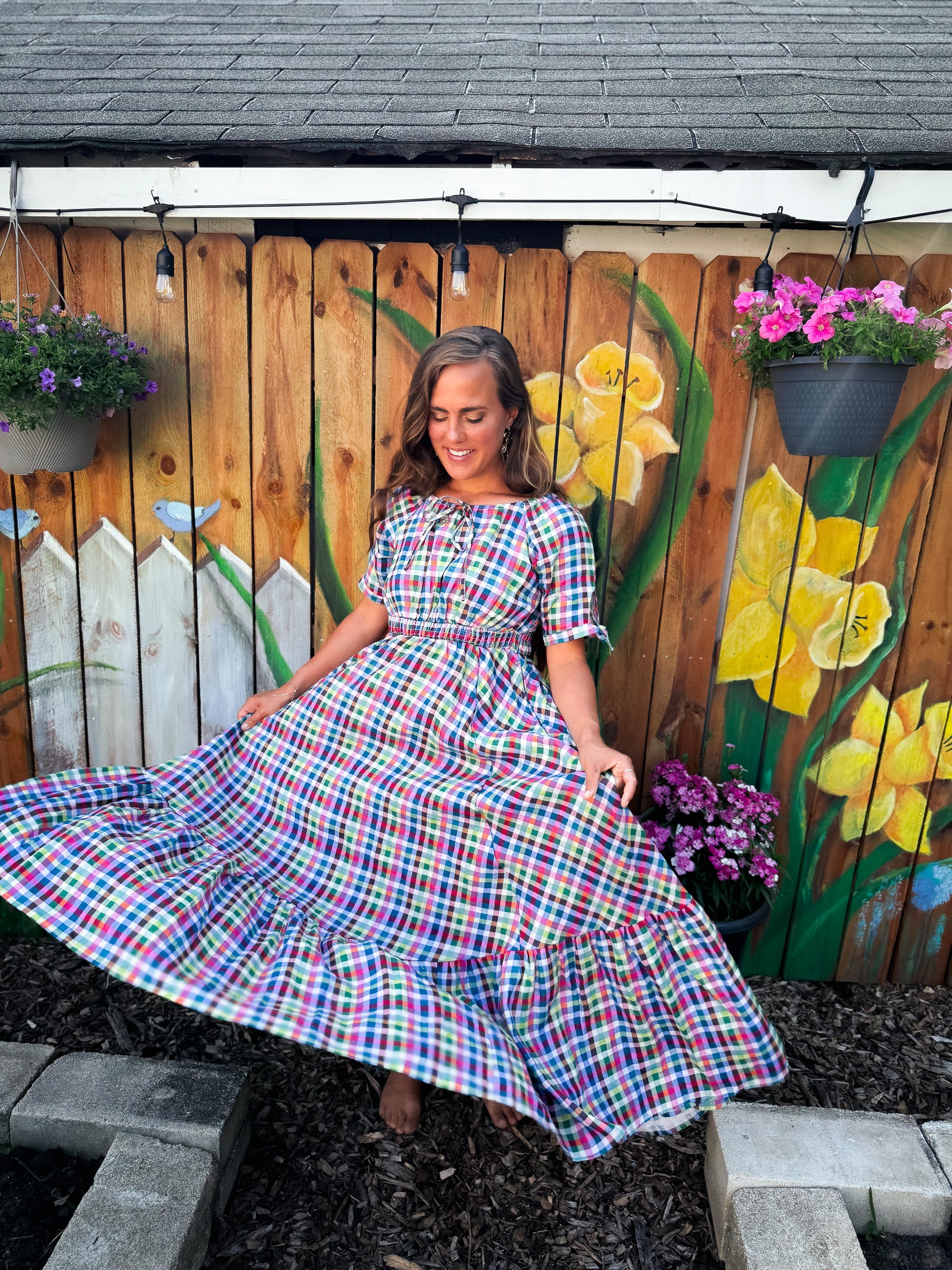 Woman in a colorful modest nursing dress standing in front of a wooden fence with floral decorations.