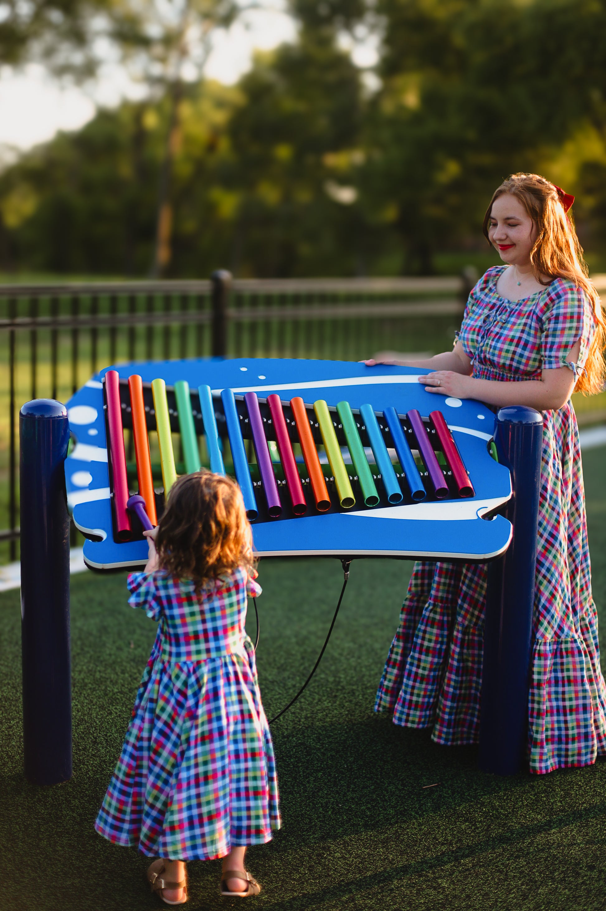 Mother and child wearing modest dresses playing with a colorful outdoor musical instrument in a park.
