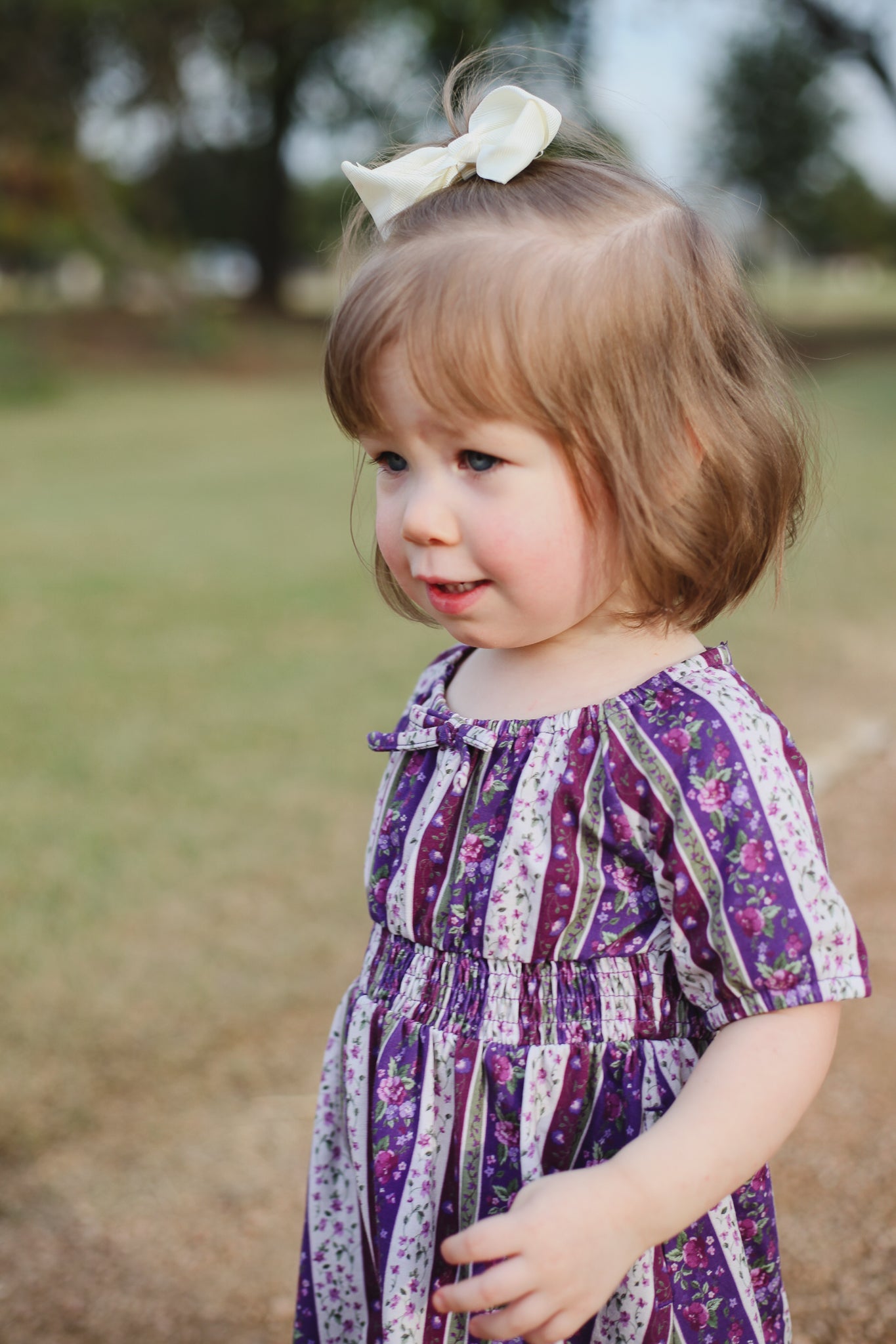 Young girl in a purple floral dress standing outdoors with a blurred natural background