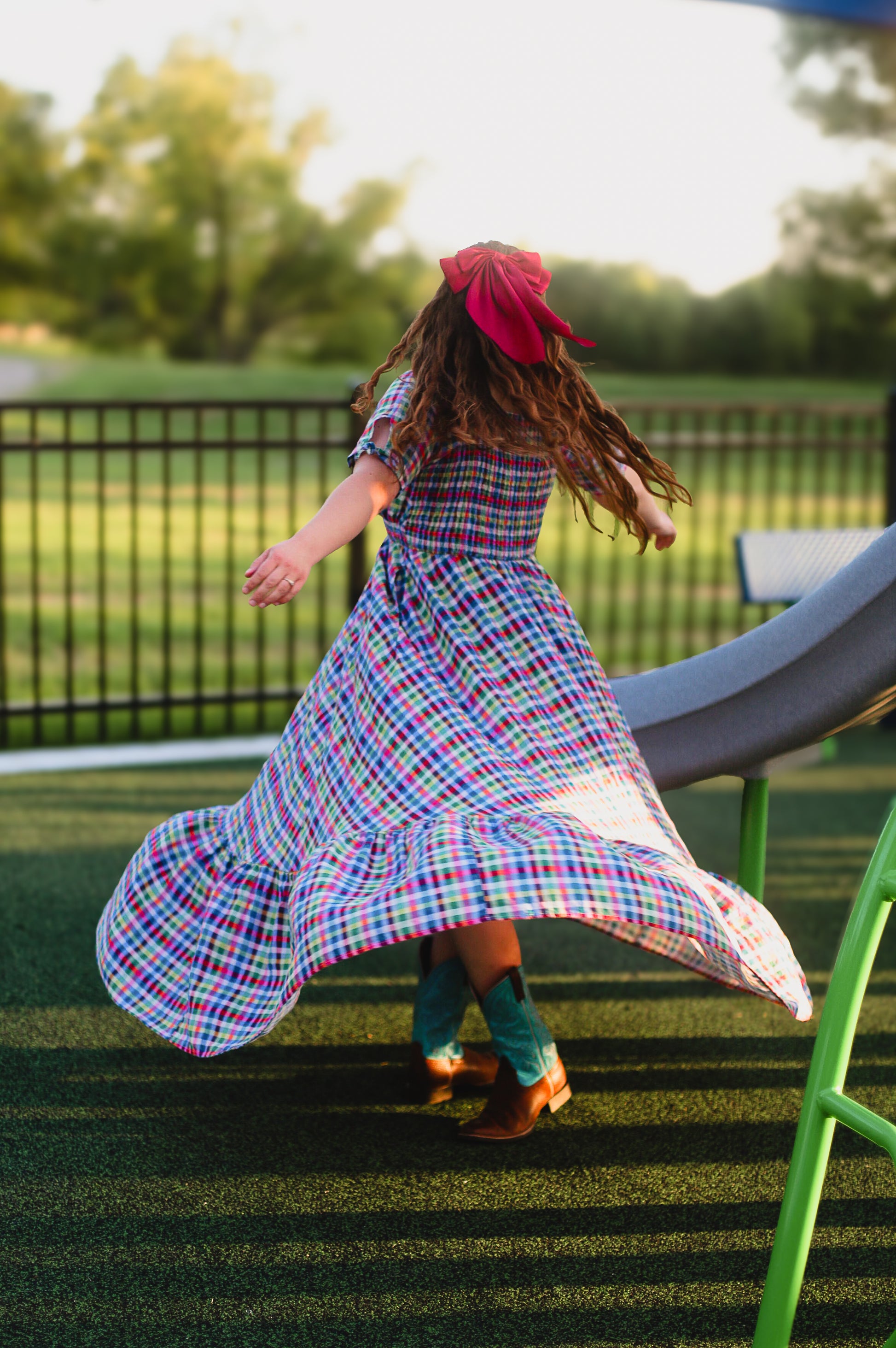 Child in a colorful modest nursing dress with a red headband running on grass with playground equipment in the background
