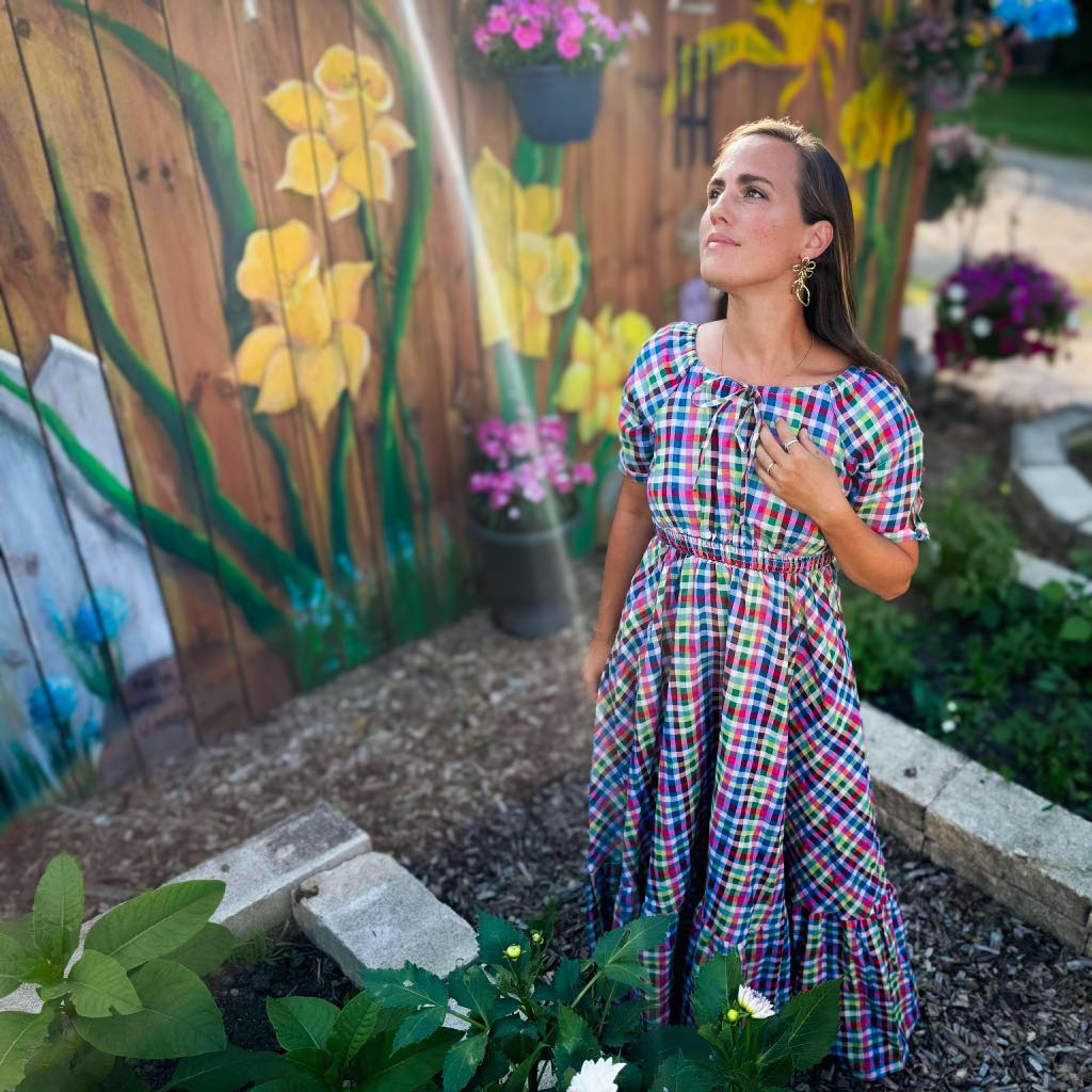 Woman in a plaid modest nursing dress standing in front of a garden with flowers and a wooden fence.