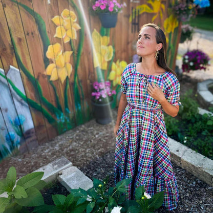 Woman in a plaid modest nursing dress standing in front of a garden with flowers and a wooden fence.