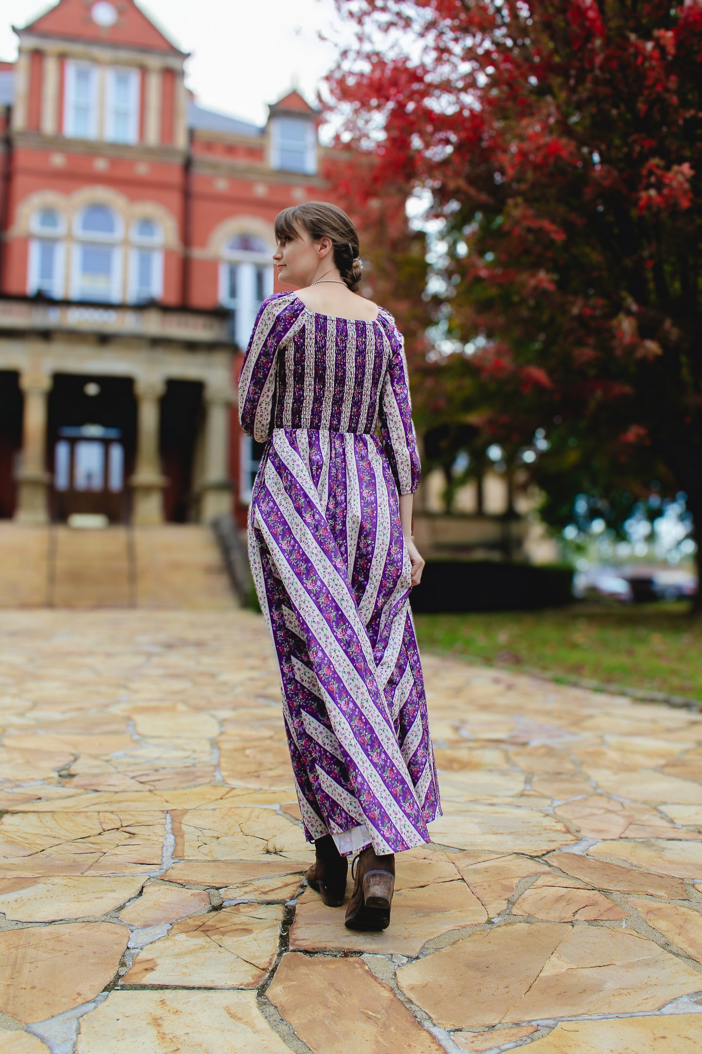 Woman in a modest nursing purple and white patterned dress standing on a stone path with a red brick building and tree in the background.