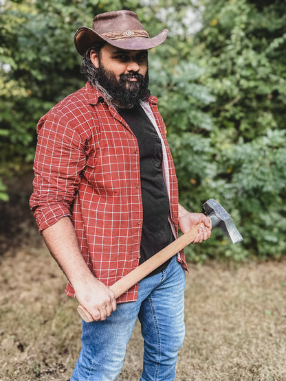 Man wearing a red plaid shirt, blue jeans, and a brown cowboy hat holding an axe outdoors.