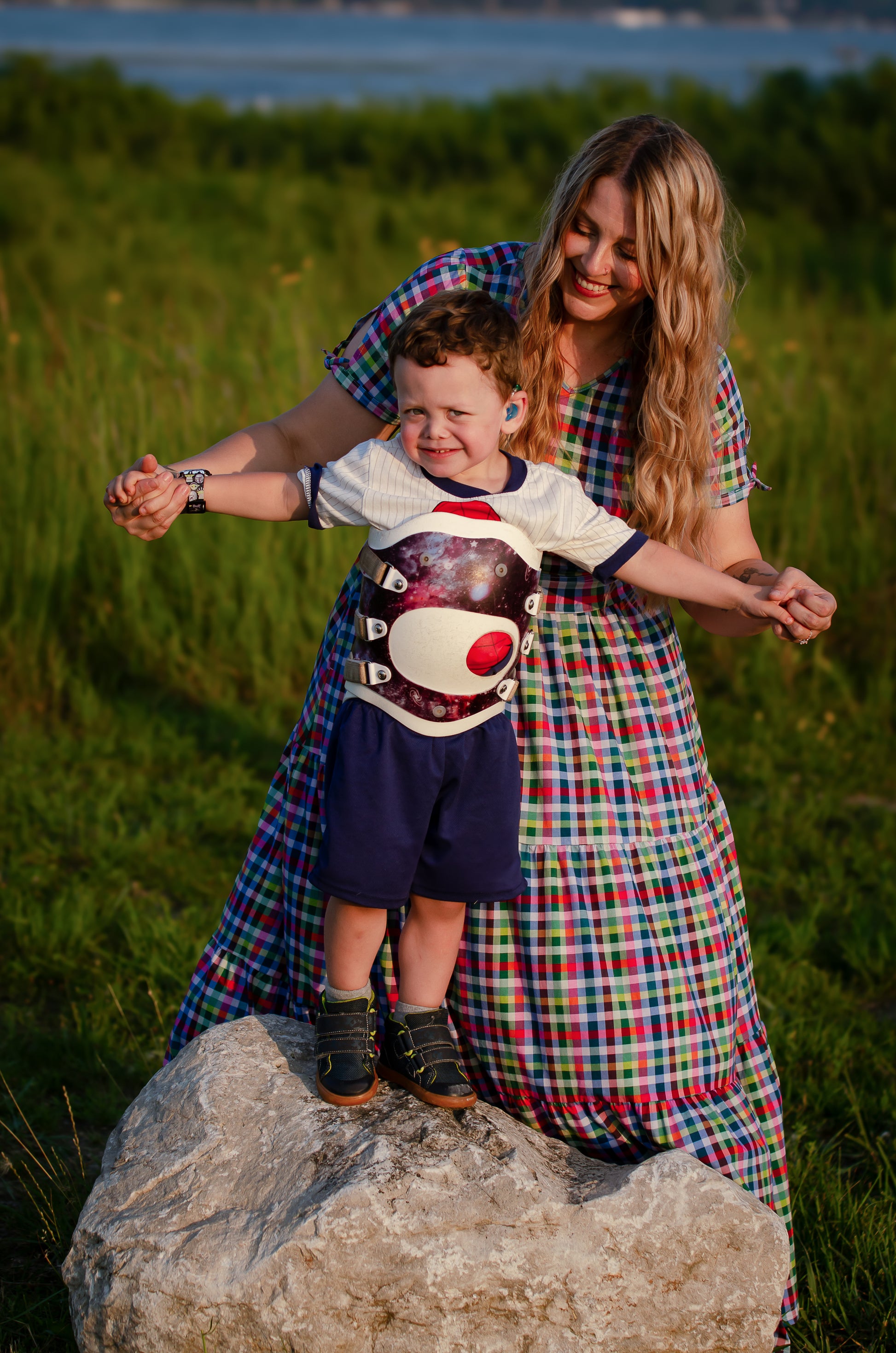 Woman in a plaid modest nursing dress holding a child on a rock with grass and water in the background