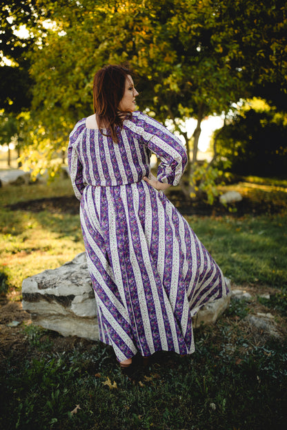 Woman in a modest nursing purple and white striped dress sitting on a rock in a natural setting.