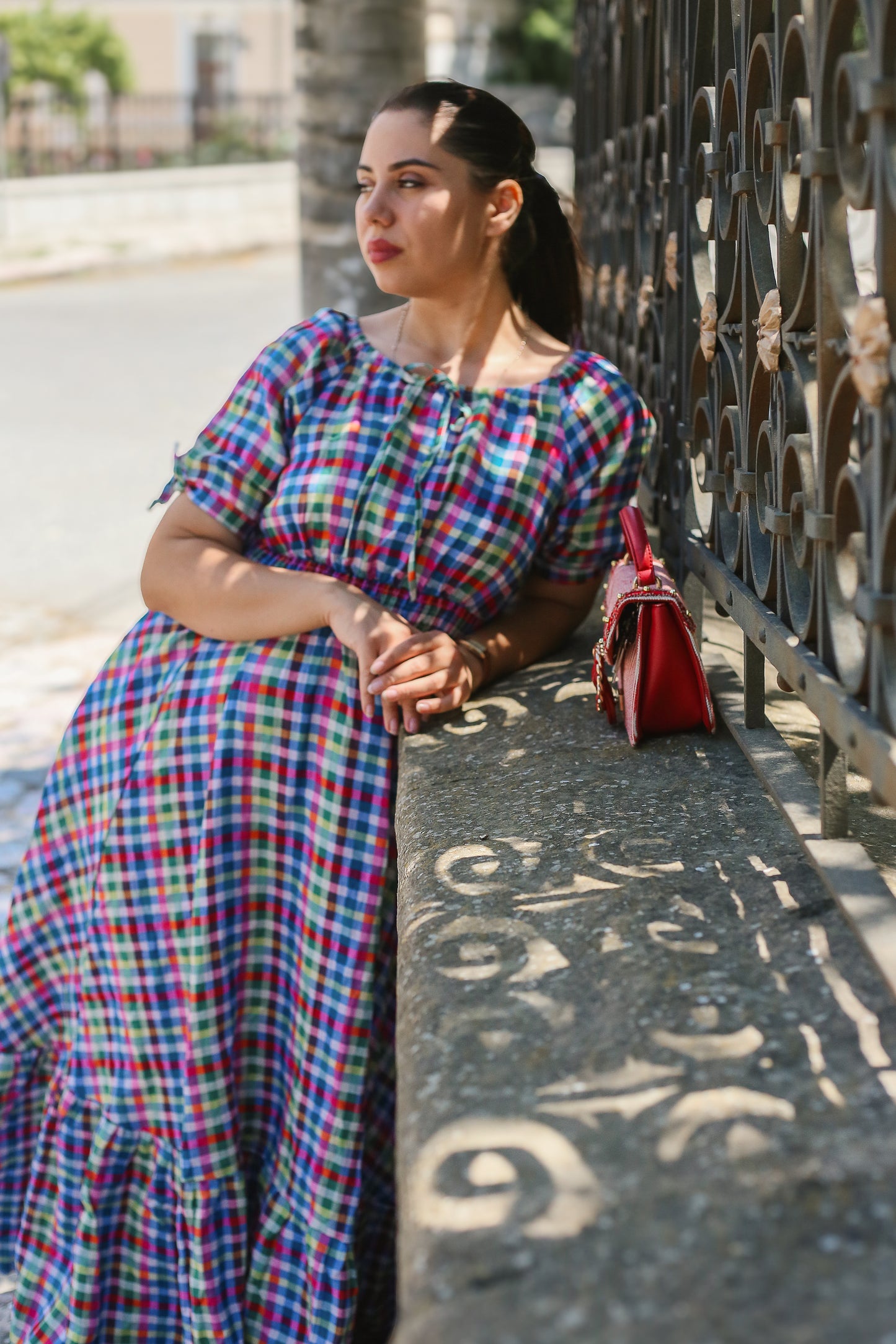 Woman in a colorful modest nursing dress leaning against a metal fence on a street.