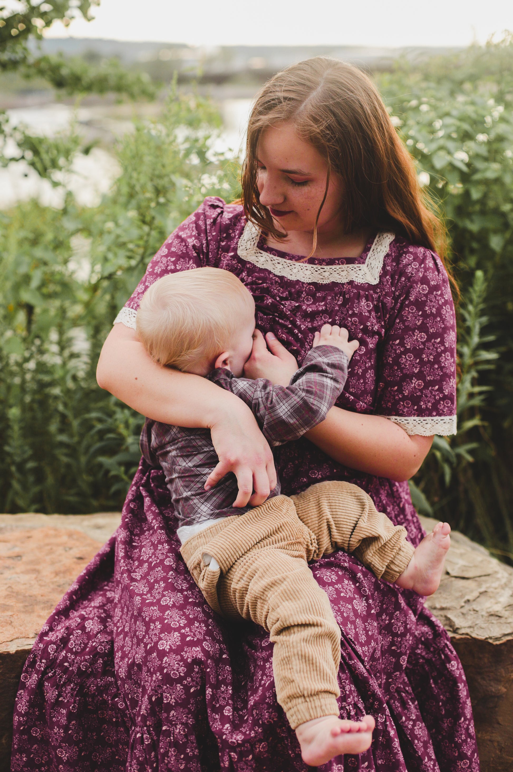 Young girl in a purple dress holding a baby outdoors with greenery in the background