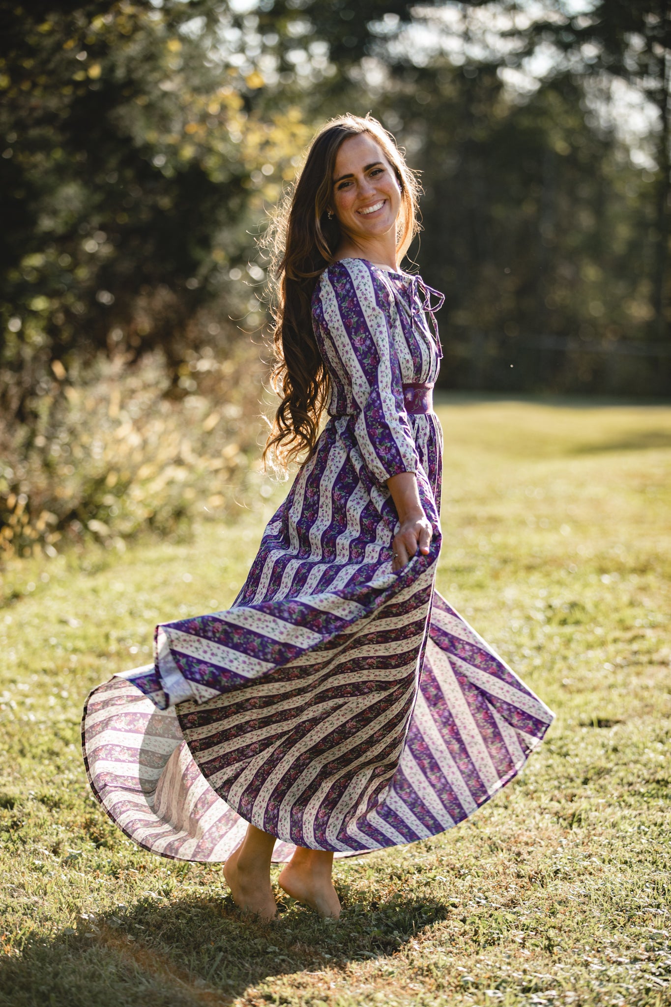Woman in a long, patterned modest nursing dress standing in a grassy field with trees in the background