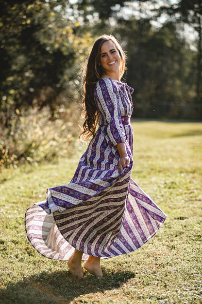 Woman in a long, patterned modest nursing dress standing in a grassy field with trees in the background