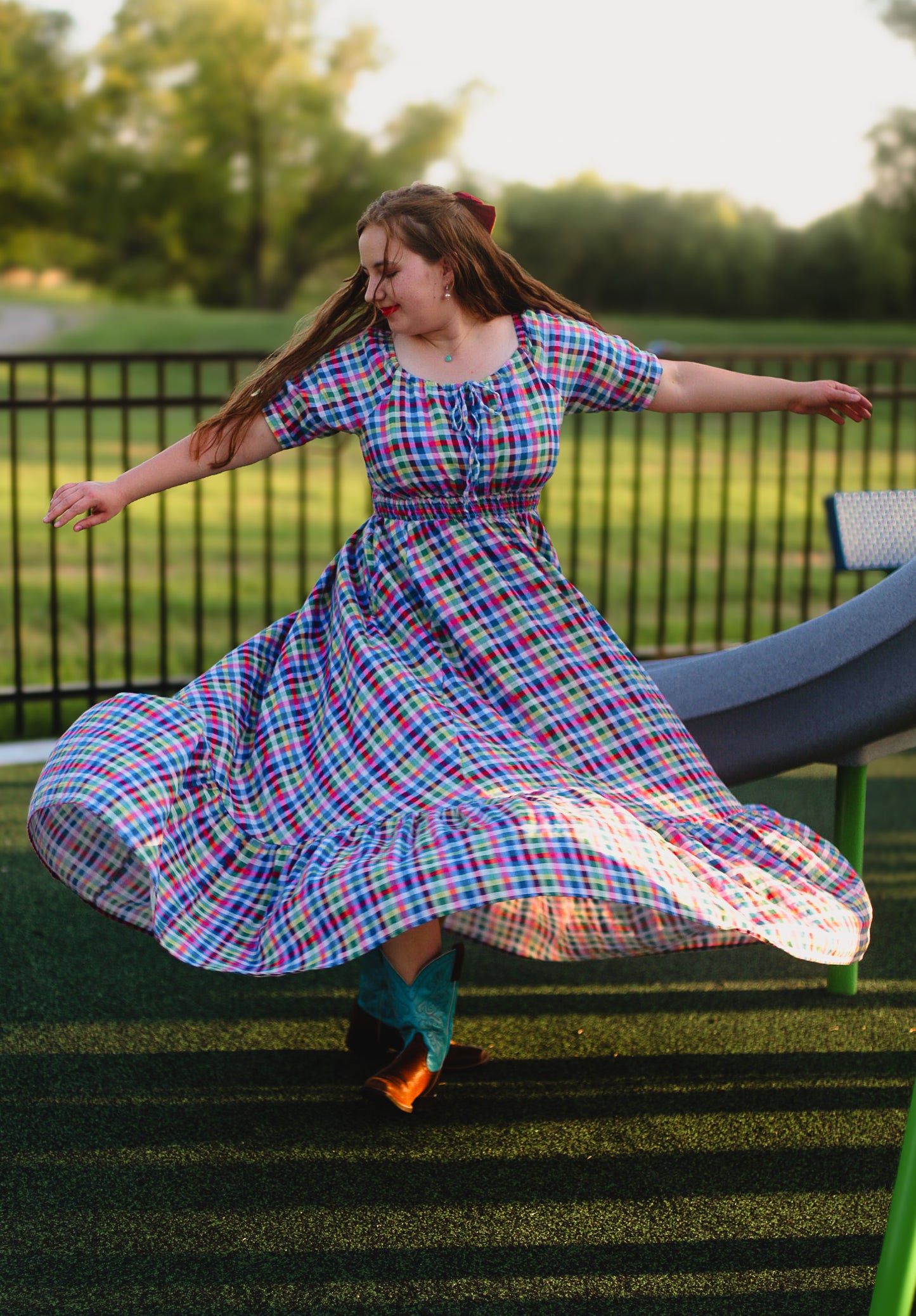 Woman in a colorful plaid modest nursing dress dancing outdoors on a grassy area with trees in the background.