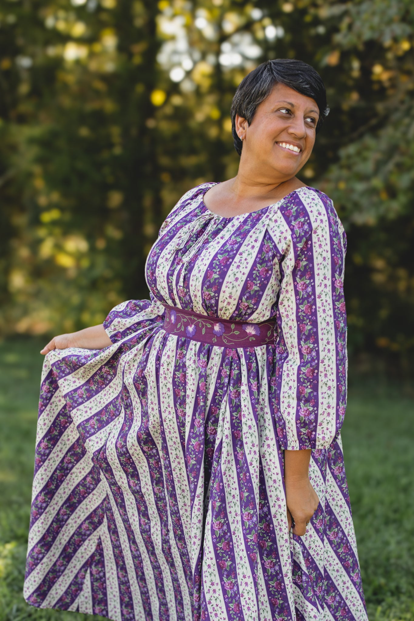 Woman wearing a modest nursing purple and white patterned dress standing outdoors with greenery in the background