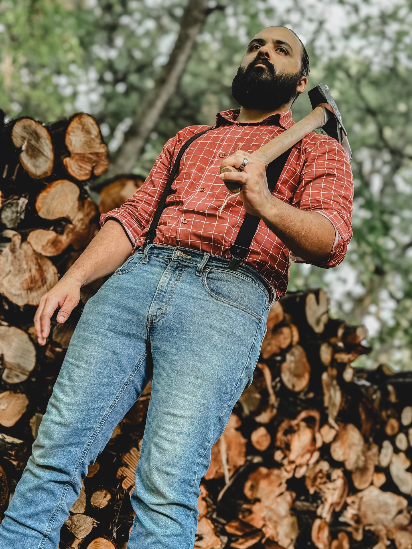 Man in red plaid shirt and blue jeans holding an axe against a background of stacked logs.
